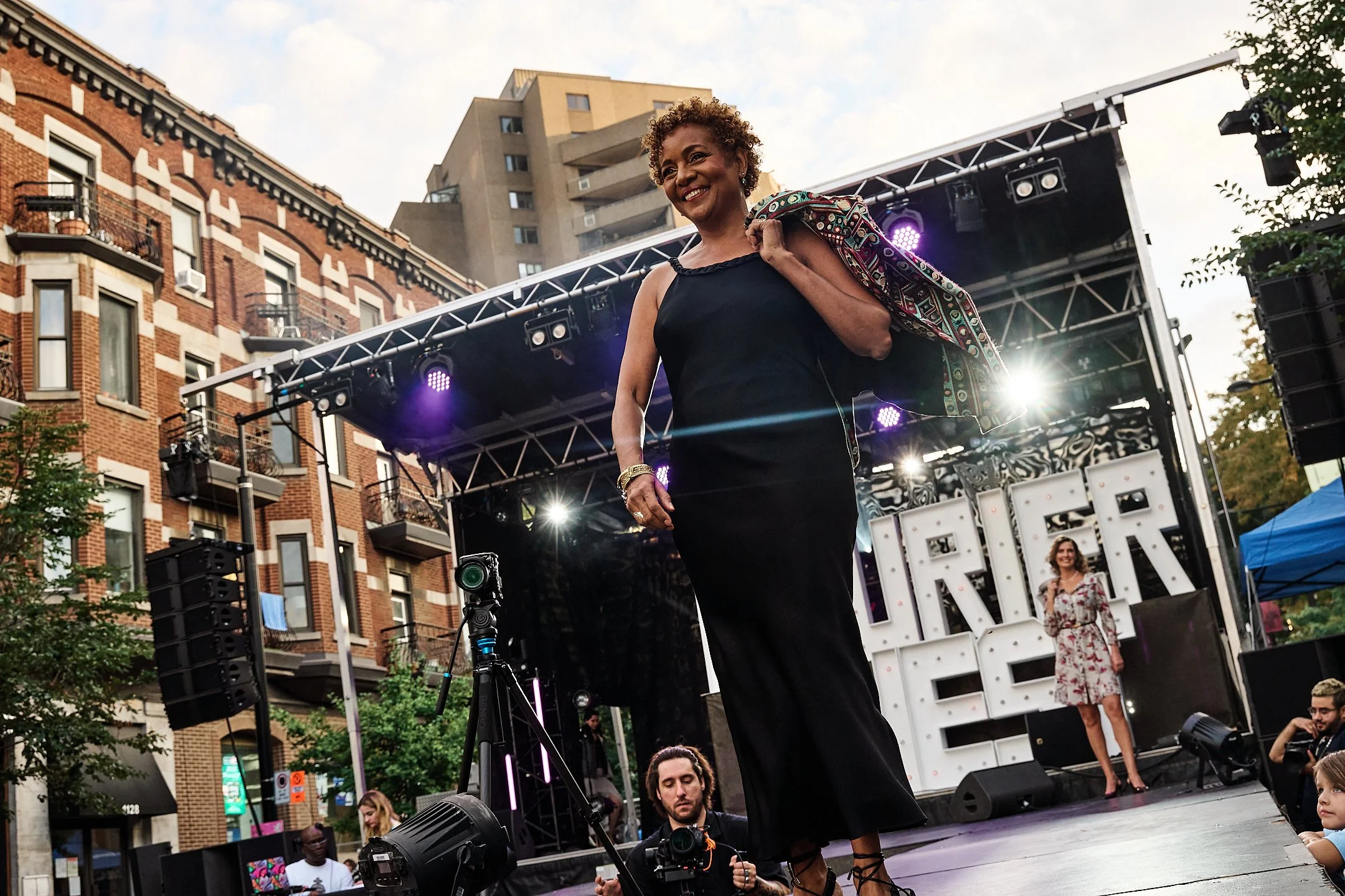 Michaëlle Jean défilant sur Laurier Ouest lors du 150ᵉ d’Outremont, vêtue d’une robe noire élégante et portant une veste colorée sur l’épaule, incarnant l’esprit festif et inclusif de la mode montréalaise.