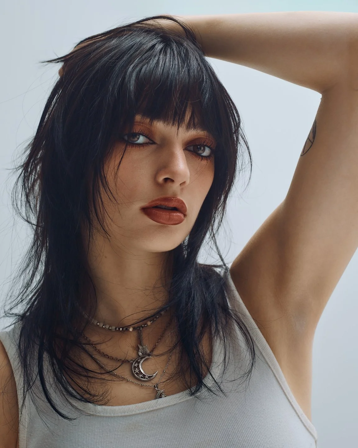 Portrait rapproché d’une mannequin en débardeur blanc, mains dans les cheveux, mettant en valeur tatouage et ceinture cloutée, shoot éditorial mode.