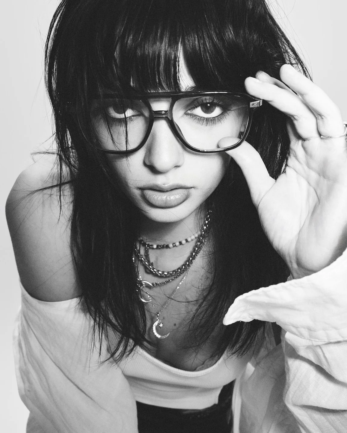 Portrait noir et blanc rapproché d’une mannequin ajustant ses lunettes oversize, stylisme minimal blanc et bijoux, shooting mode attitude.