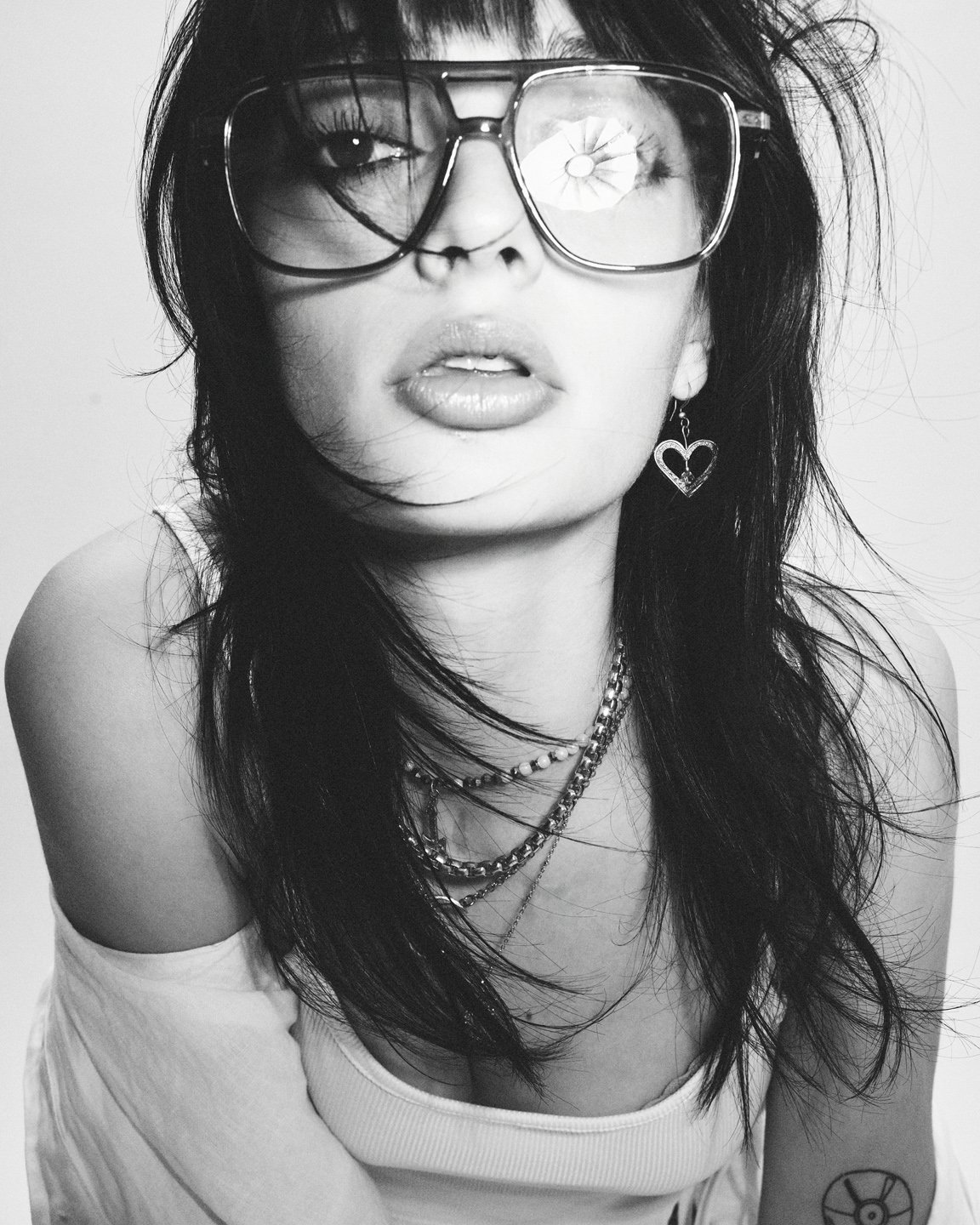 Portrait noir et blanc d’une mannequin portant de grandes lunettes transparentes, cheveux décoiffés et colliers superposés, photographie éditoriale mode.