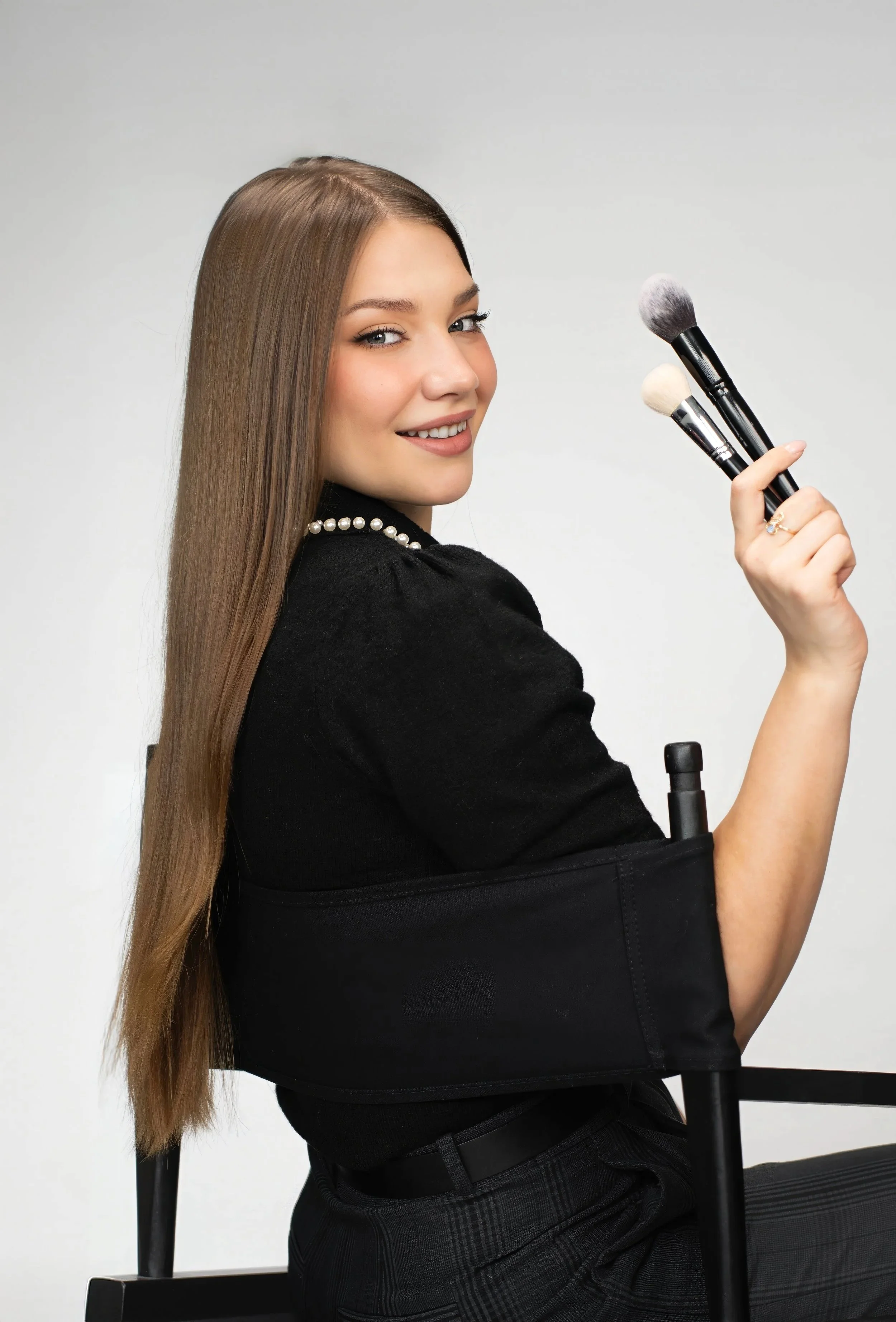 Toronto's trusted bridal makeup artist Masha Mez is featured sitting in a makeup chair holding up 3 makeup brushes and warmly smiling at the camera.
