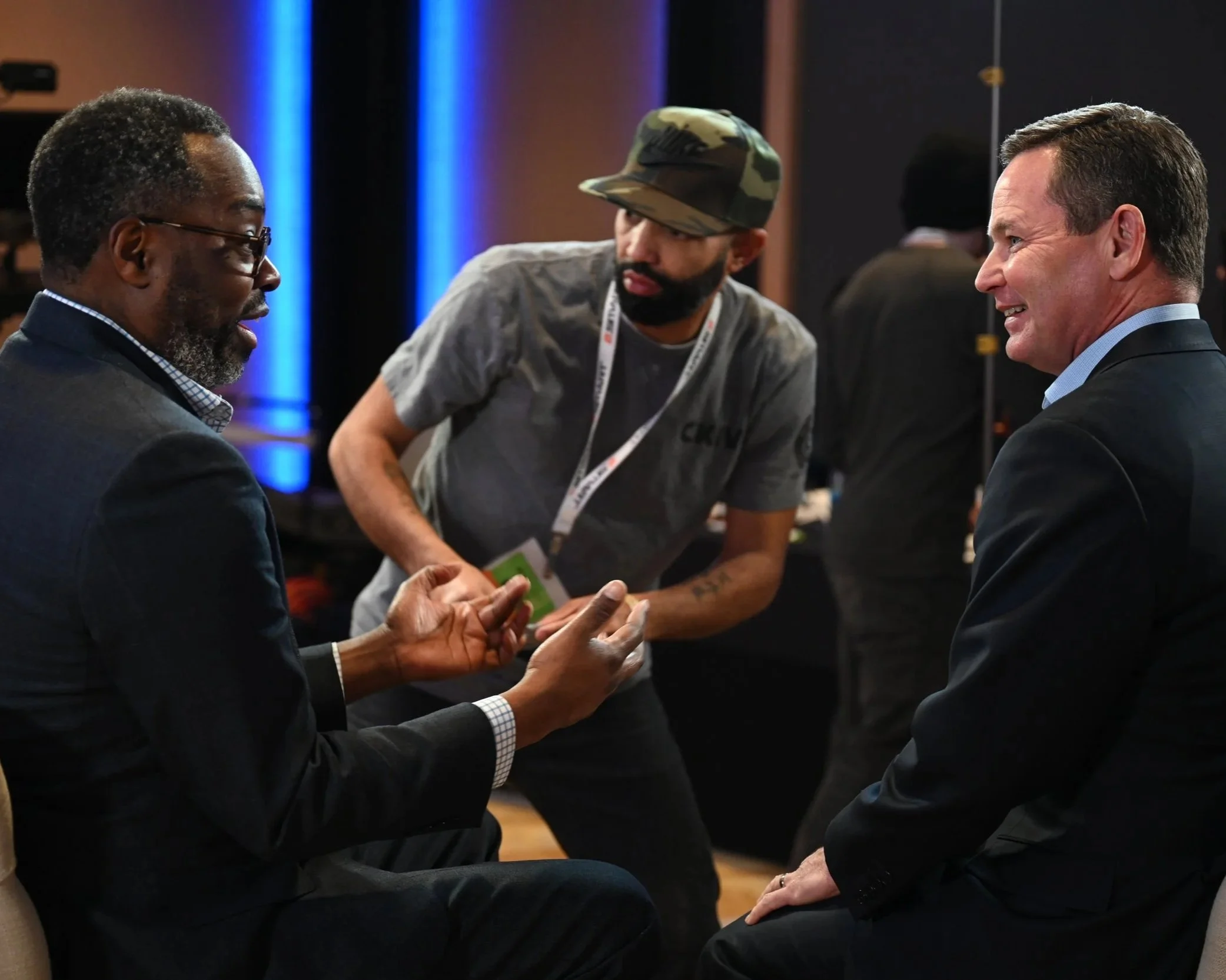 Three men engaged in conversation, with one wearing a navy suit and glasses, another wearing a cap and casual grey T-shirt, and the third in a dark suit with a blue shirt, indoors with blue lighting in the background.