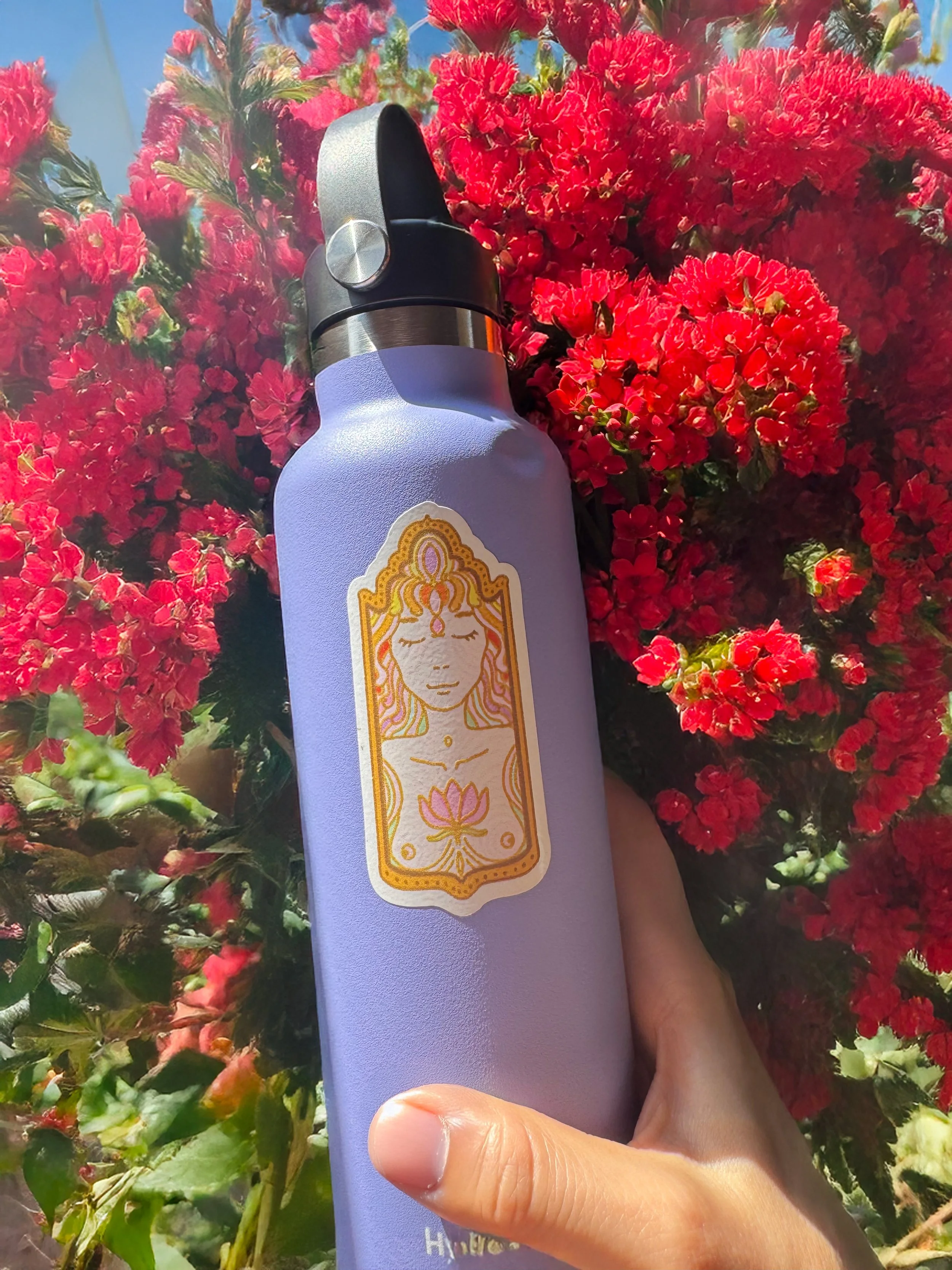 A lavender-colored spray bottle with a decorative label depicting a peaceful meditative woman with closed eyes, surrounded by carved flowers, held in front of vibrant red flowering plants under a blue sky.