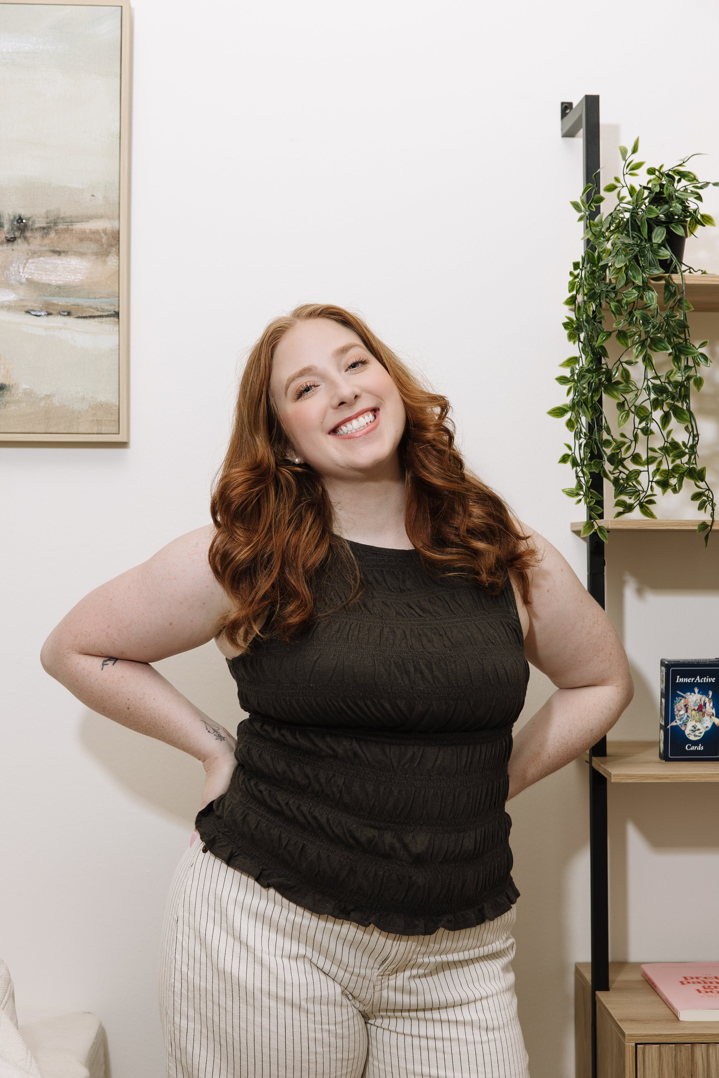 A smiling woman with red hair and tattoos on her arms stands in a room with white walls, a painting, some green plants, and books on a shelf.