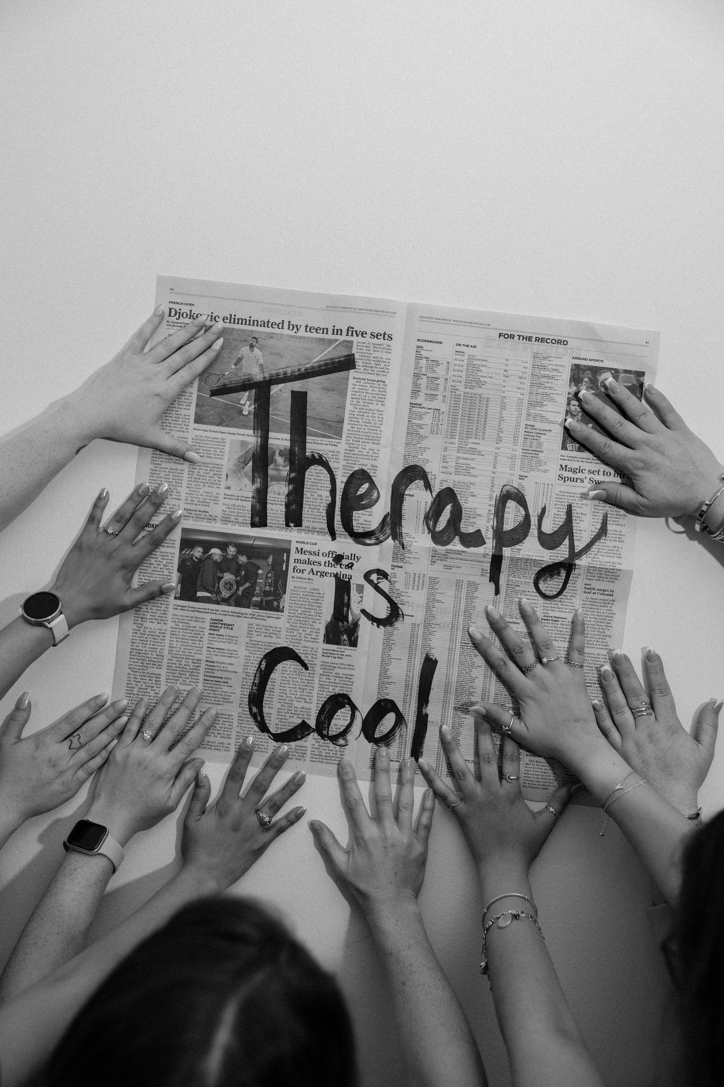 Multiple hands of diverse people reaching toward a newspaper on a table, with the words 'Therapy is Cool' written across it in black ink.