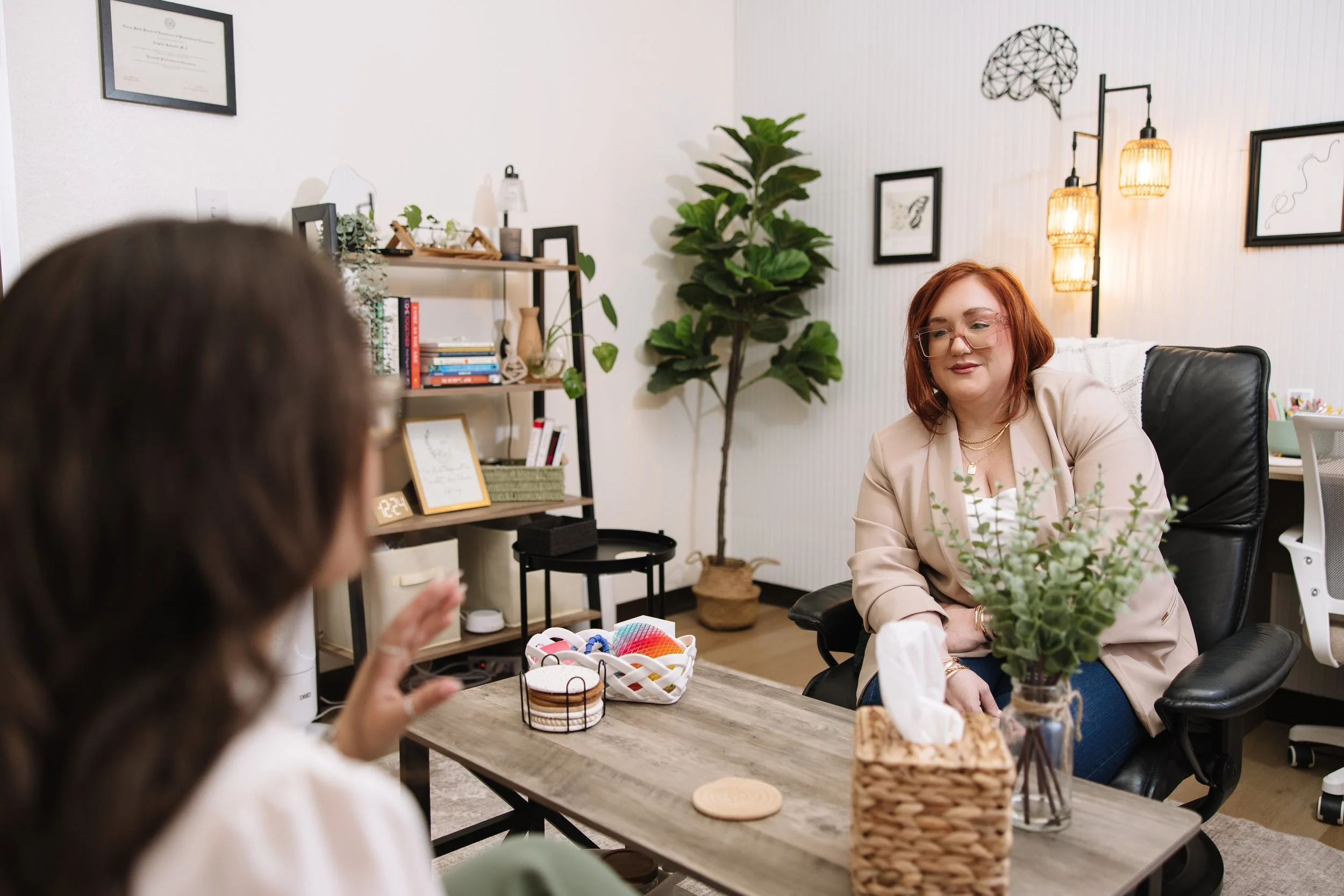 A woman with red hair and glasses sitting at a table, engaged in conversation with another person whose face is partially visible. The setting is a cozy, well-decorated office or lounge area with framed pictures, a large plant, and a bookshelf in the background. The table has a basket of craft supplies, and there is a small clock and a framed document on the shelf.