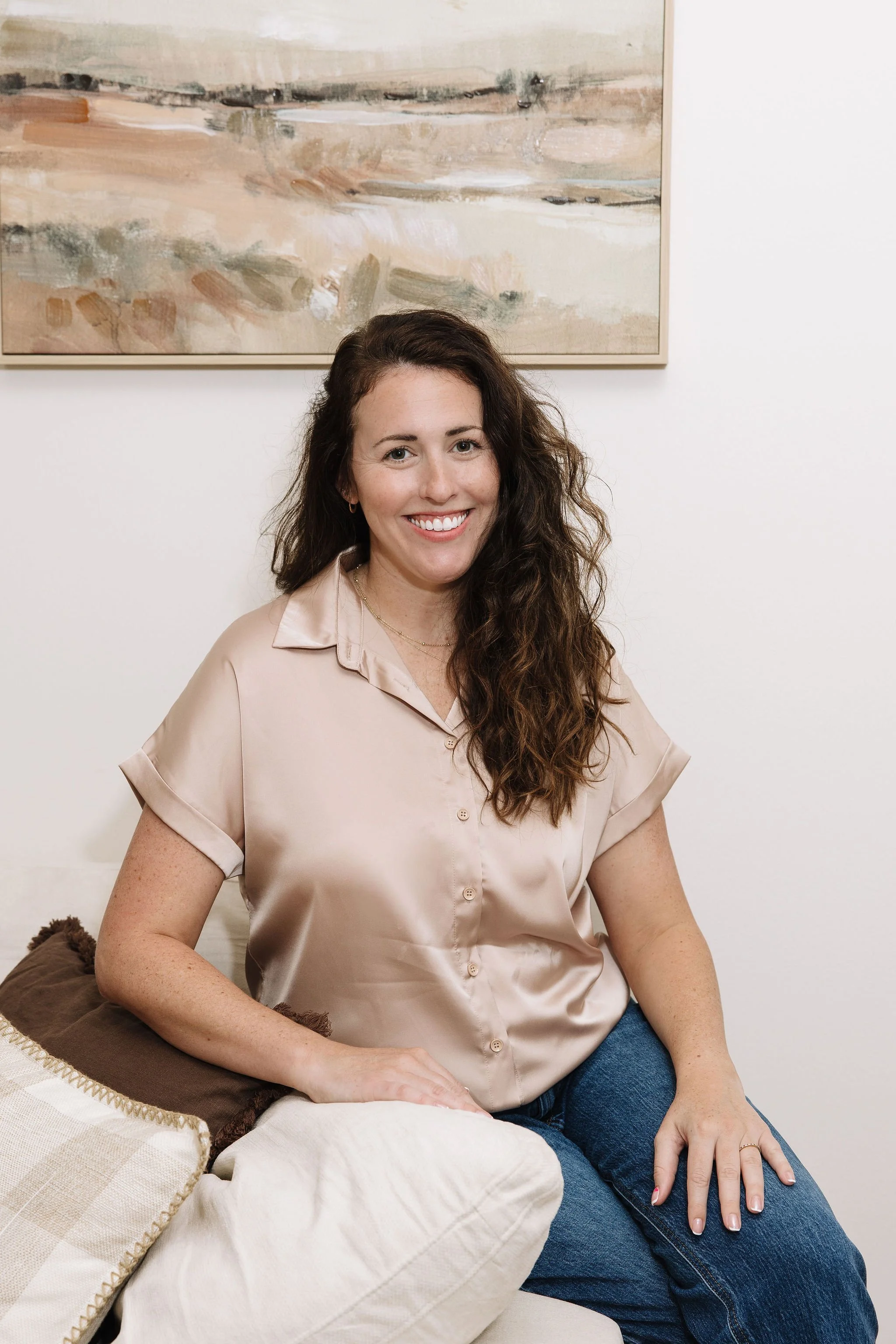 A woman with long, curly brown hair, wearing a red polka-dot blouse, a black jacket, and blue jeans, standing against a brick wall, smiling at the camera.