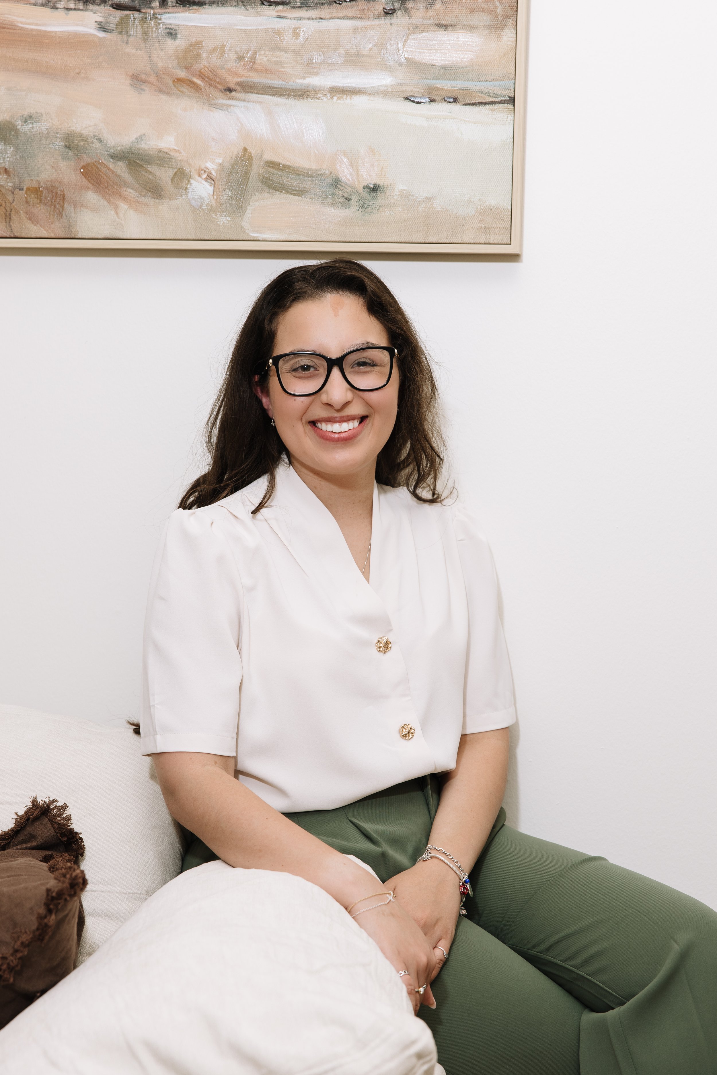 A woman with long dark hair, glasses, and a wide smile sitting on a white couch in front of a plain white wall and a framed abstract painting, wearing a white blouse and green pants.