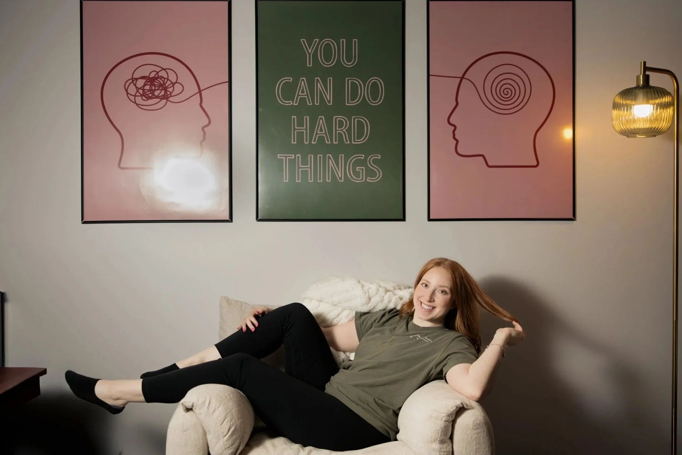 A woman with red hair is lying on a beige armchair, smiling and holding her hair with one hand while resting her other arm on the armrest. Behind her, there are three framed art pieces on a gray wall, with the middle one displaying the phrase 'You Can Do Hard Things' in white text on a green background. A gold floor lamp with a ribbed glass shade is on the right side of the image.