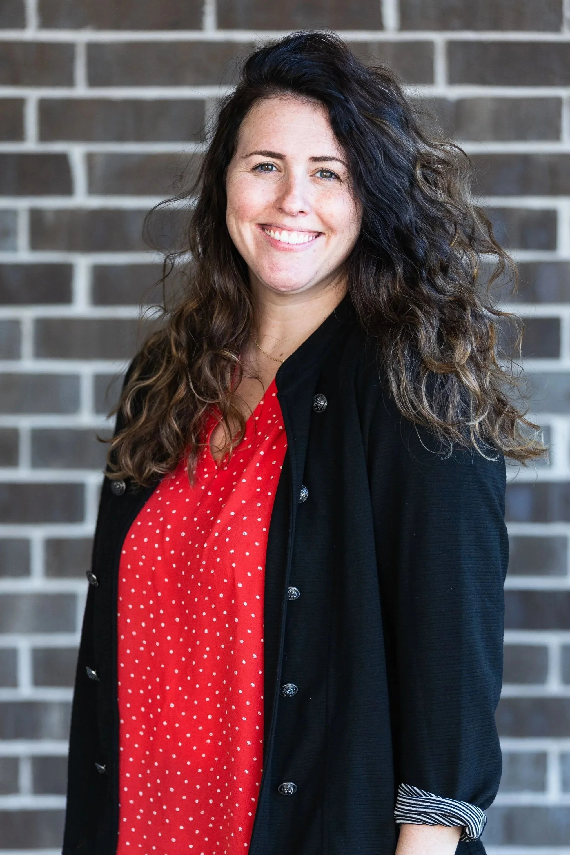 A woman with long, curly dark hair and fair skin, smiling, wearing a red dress with white polka dots and a black cardigan, standing in front of a brick wall.