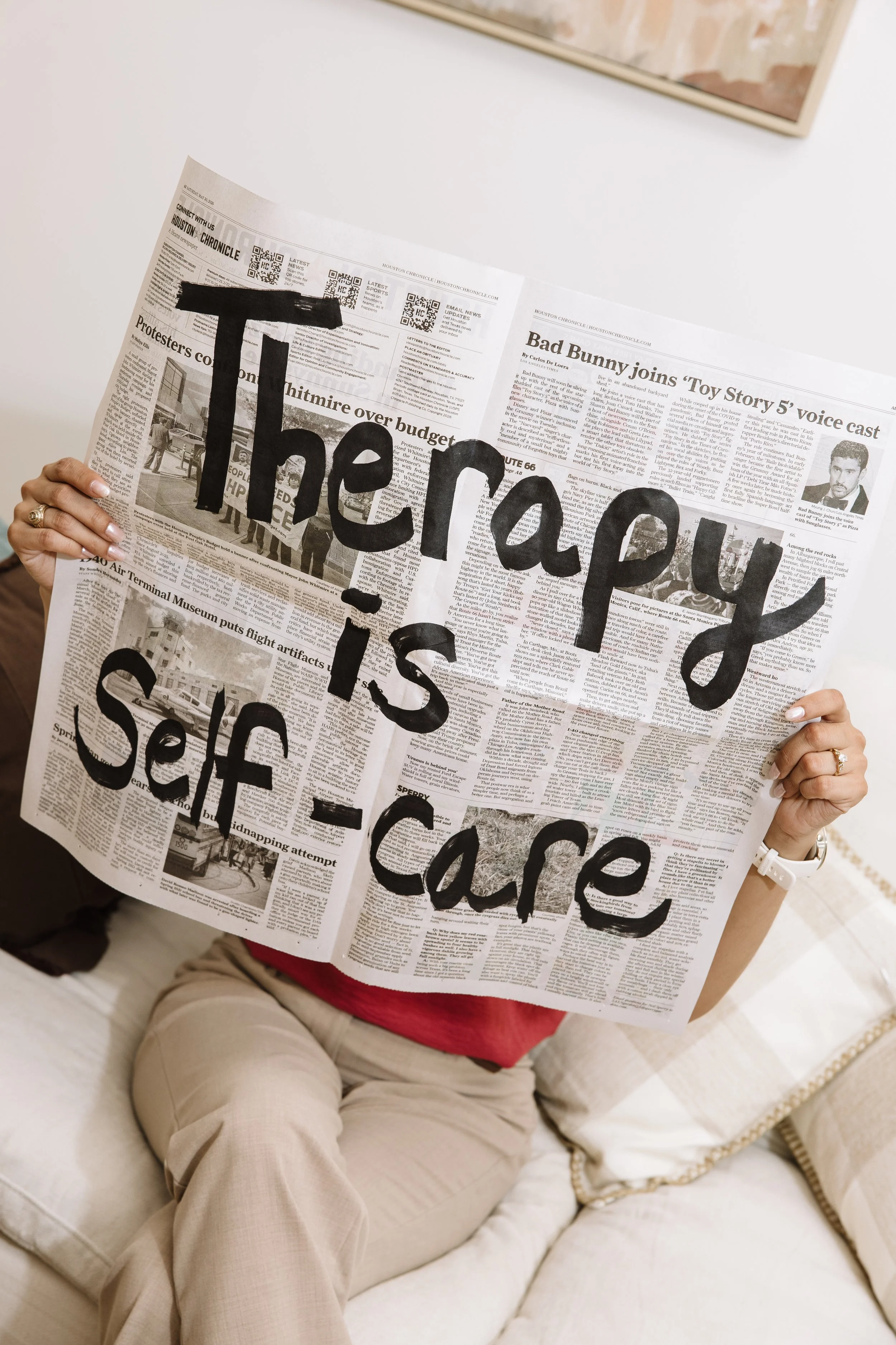 Person sitting on a beige sofa, holding a newspaper with large handwritten-style black text covering the front page.