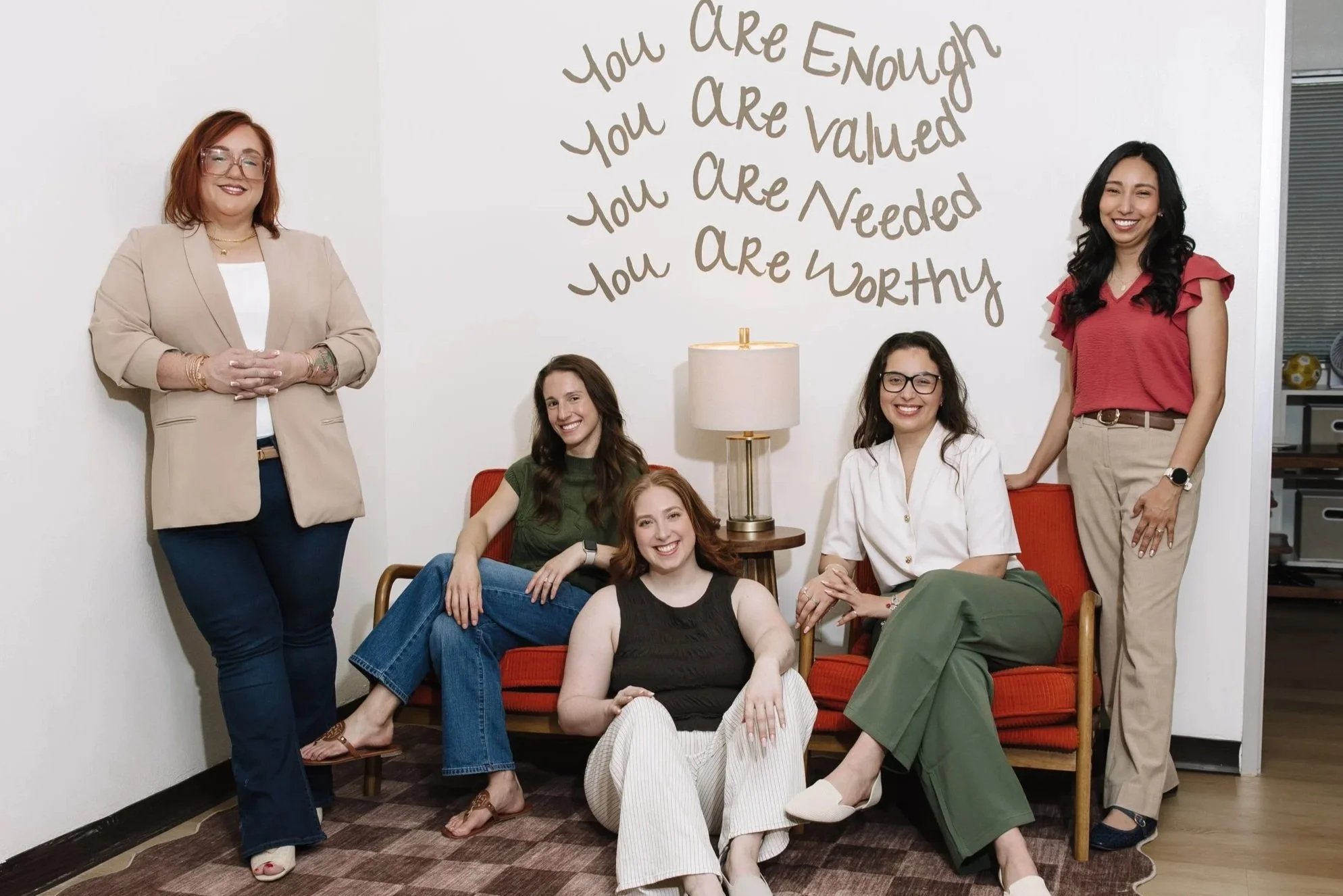 Five women posing in a living room with a wall decoration that reads, "You are enough, you are valued, you are needed, you are worthy."