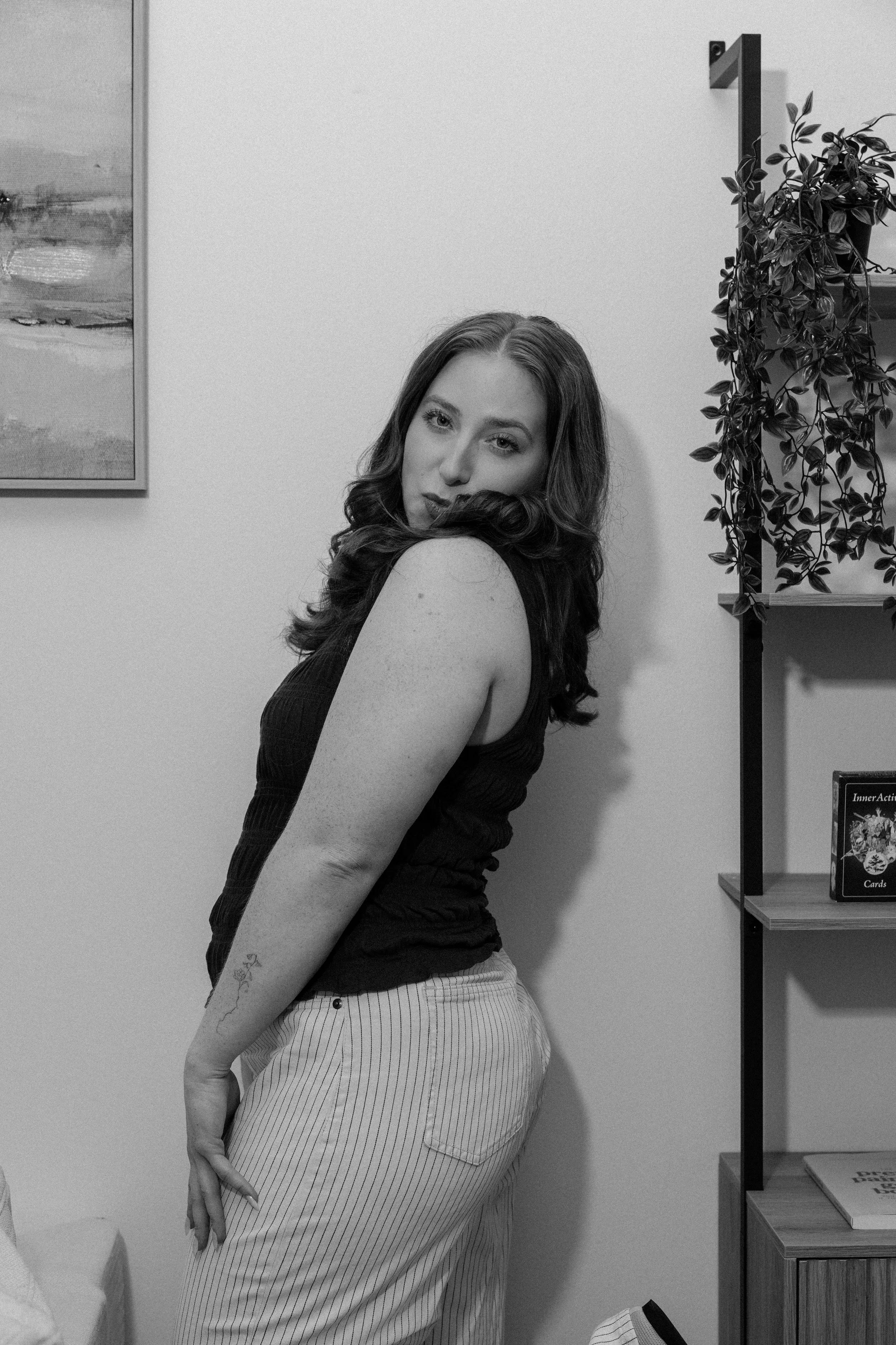 A woman with wavy hair wearing a sleeveless top and striped pants standing in a room with bookshelves, plants, and artwork on the wall.
