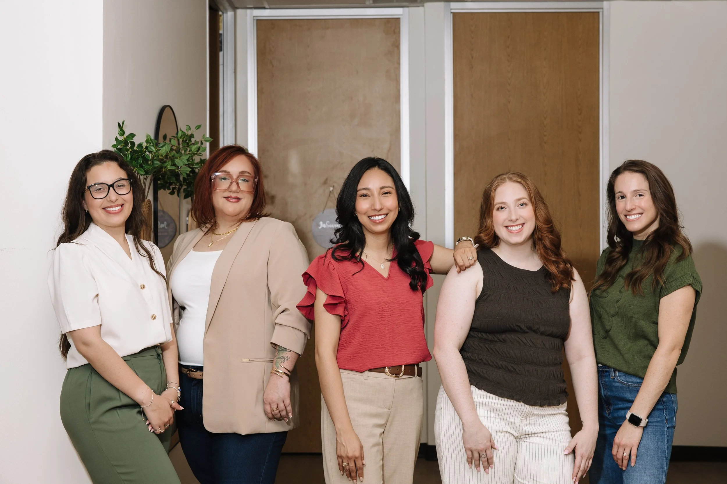 Group of five women standing together in an indoor setting, smiling, wearing casual and business casual attire.