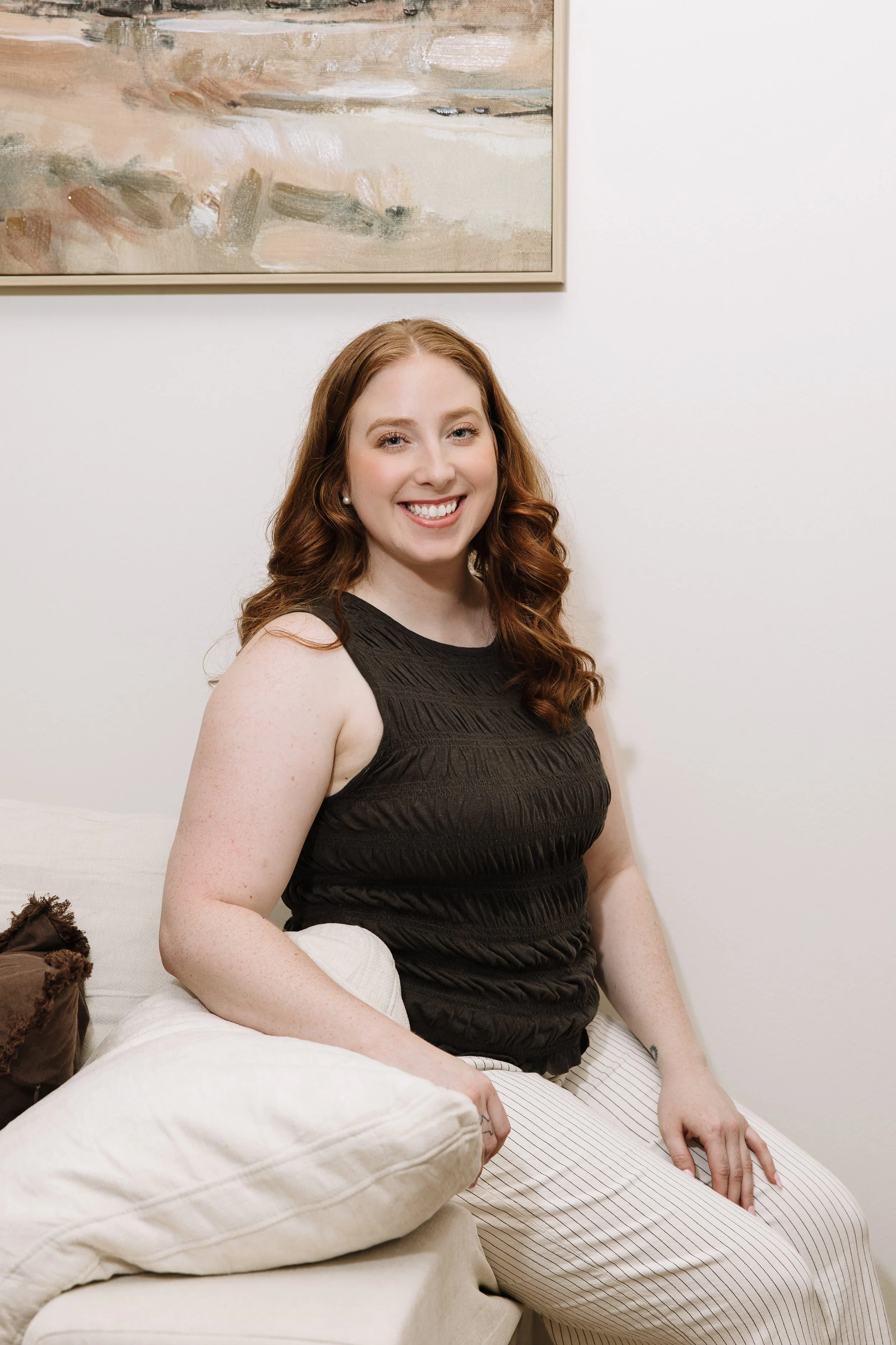 Woman, Red Headed Therapist sitting on therapy couch with hands crossed in front of her with warm welcoming smile