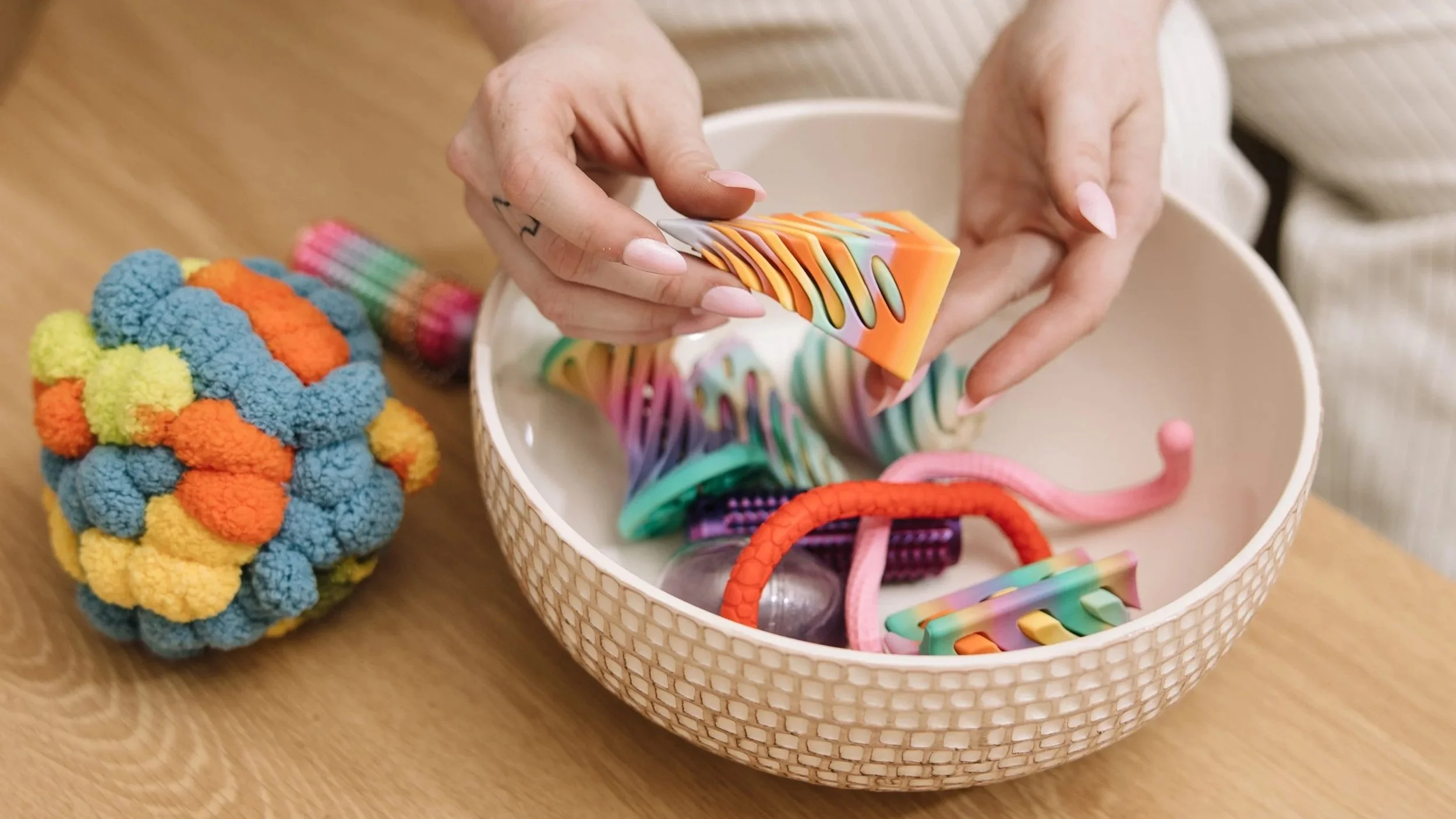 Hands holding a colorful soap in front of a basket of assorted rainbow-colored hair accessories and a crocheted rainbow plush toy on a wooden table.