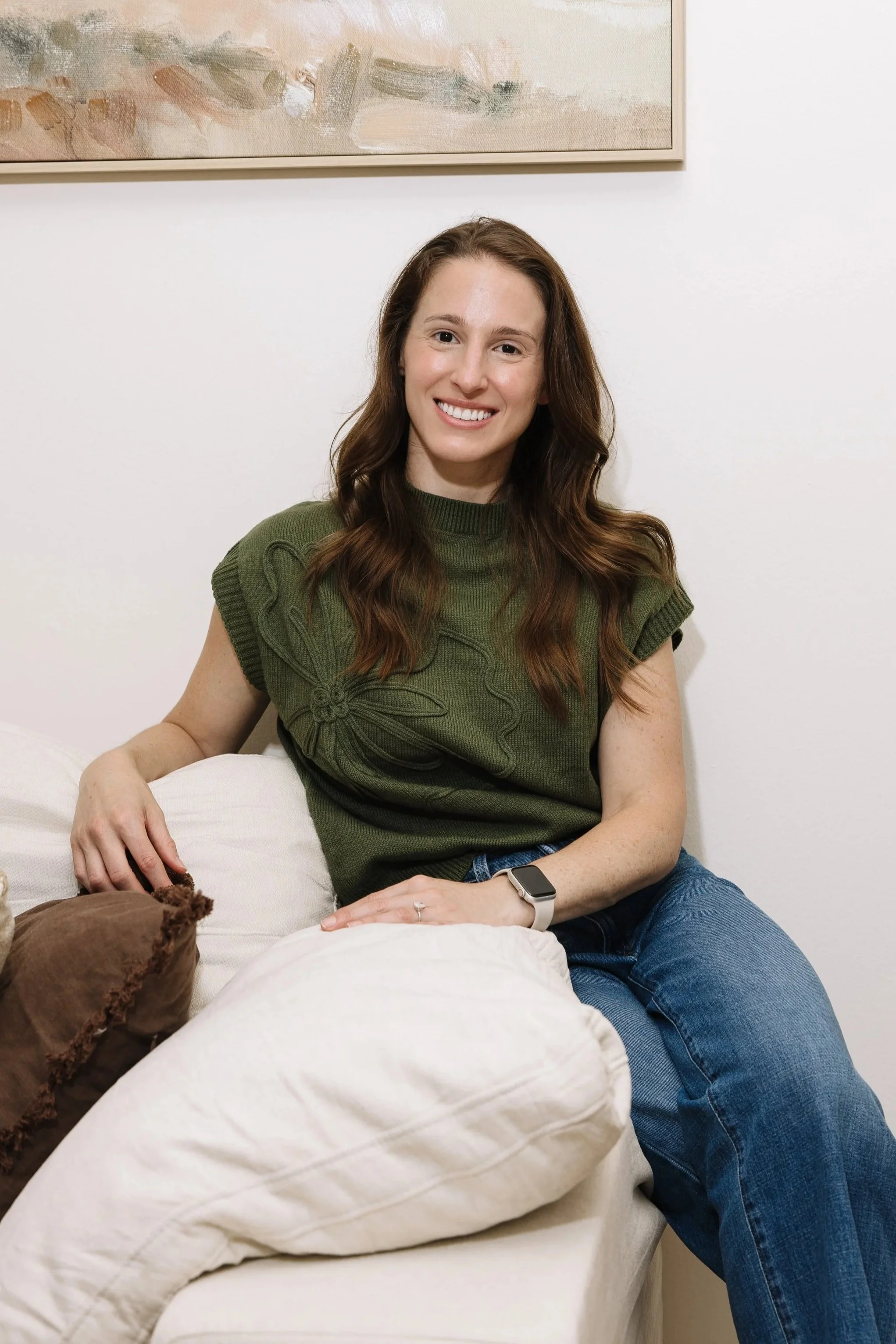 A woman with long brown hair and a smile, wearing a green sleeveless sweater and blue jeans, sitting on a cream-colored couch in a room with white walls and a framed abstract painting.