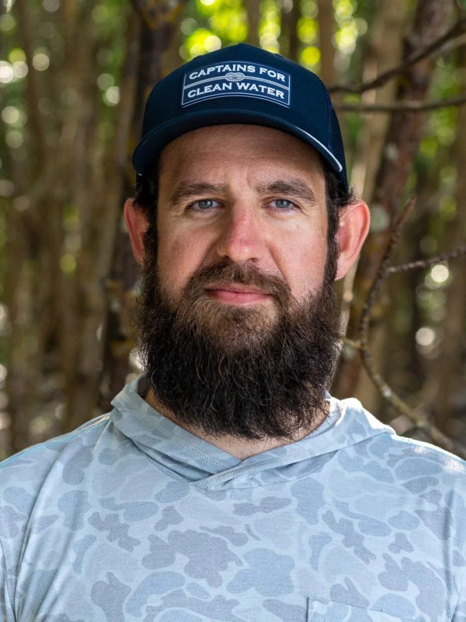 A man with a beard wearing a blue cap that says "Captains for Clean Water" and a light-colored, patterned athletic shirt, standing outdoors with blurred trees in the background.