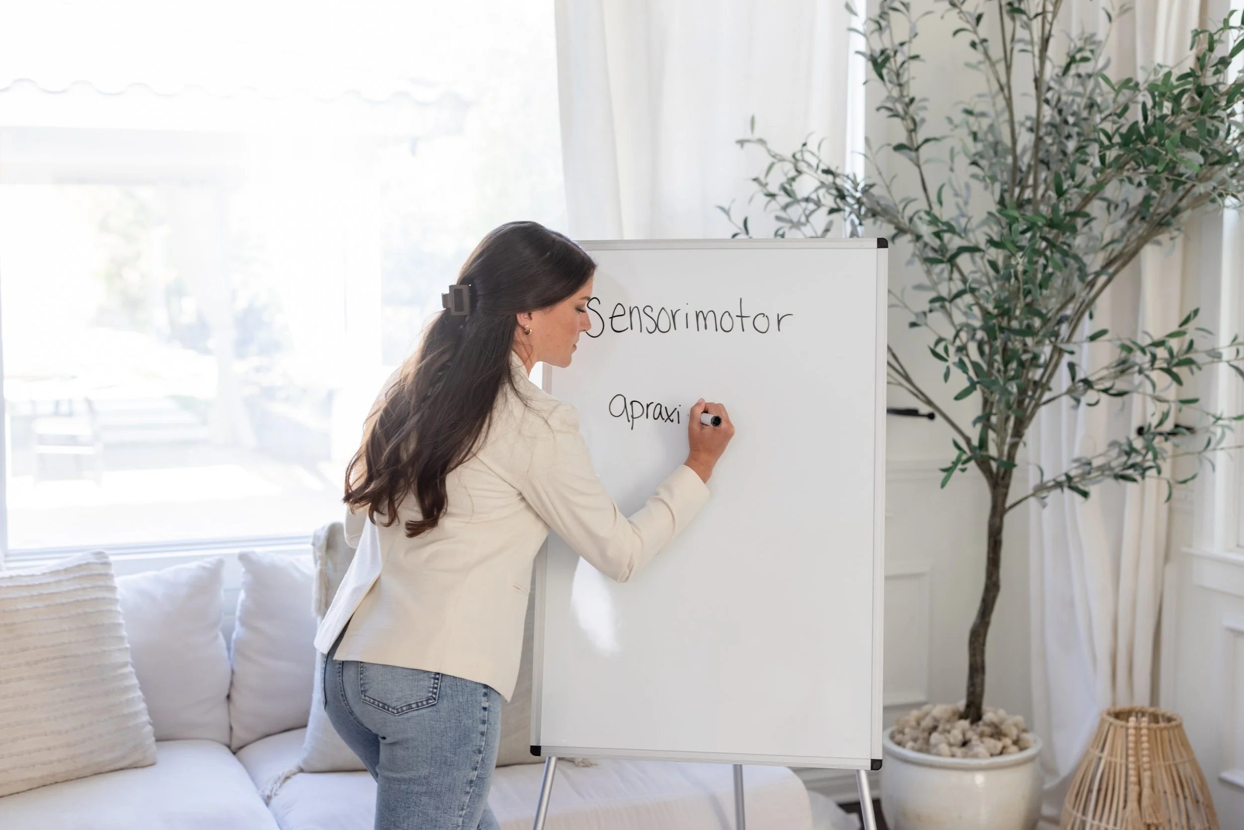 Erin Clarelli, Autism Therapist, wearing a crisp white blazer and jeans writes the word "sensorimotor" on a whiteboard.