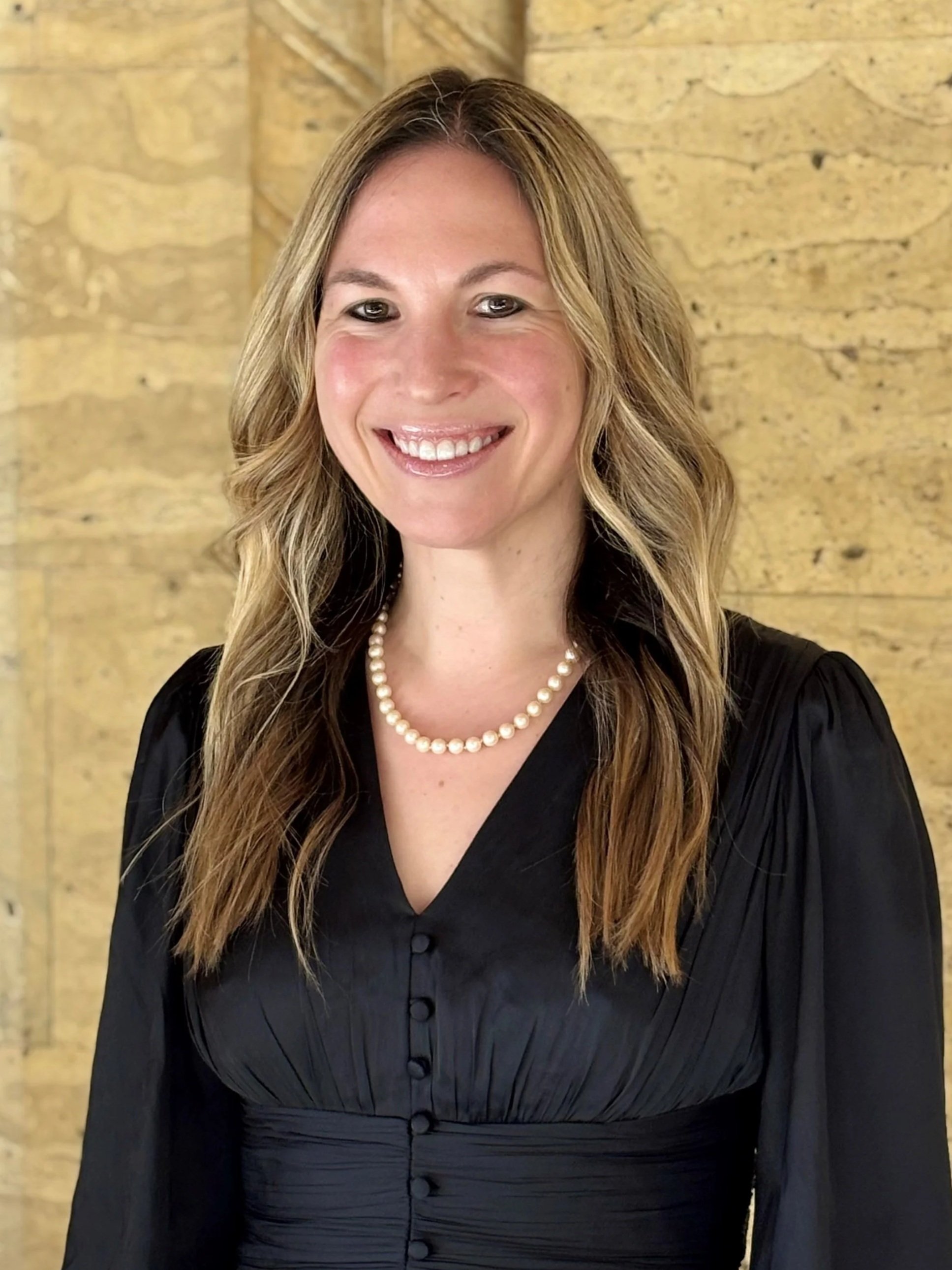 Headshot of FemmeFactor Expert, Helena Rosenthal a Financial Advisor.  She is wearing a black dress with buttons and a pearl necklace, smiling in front of a wooden wall.