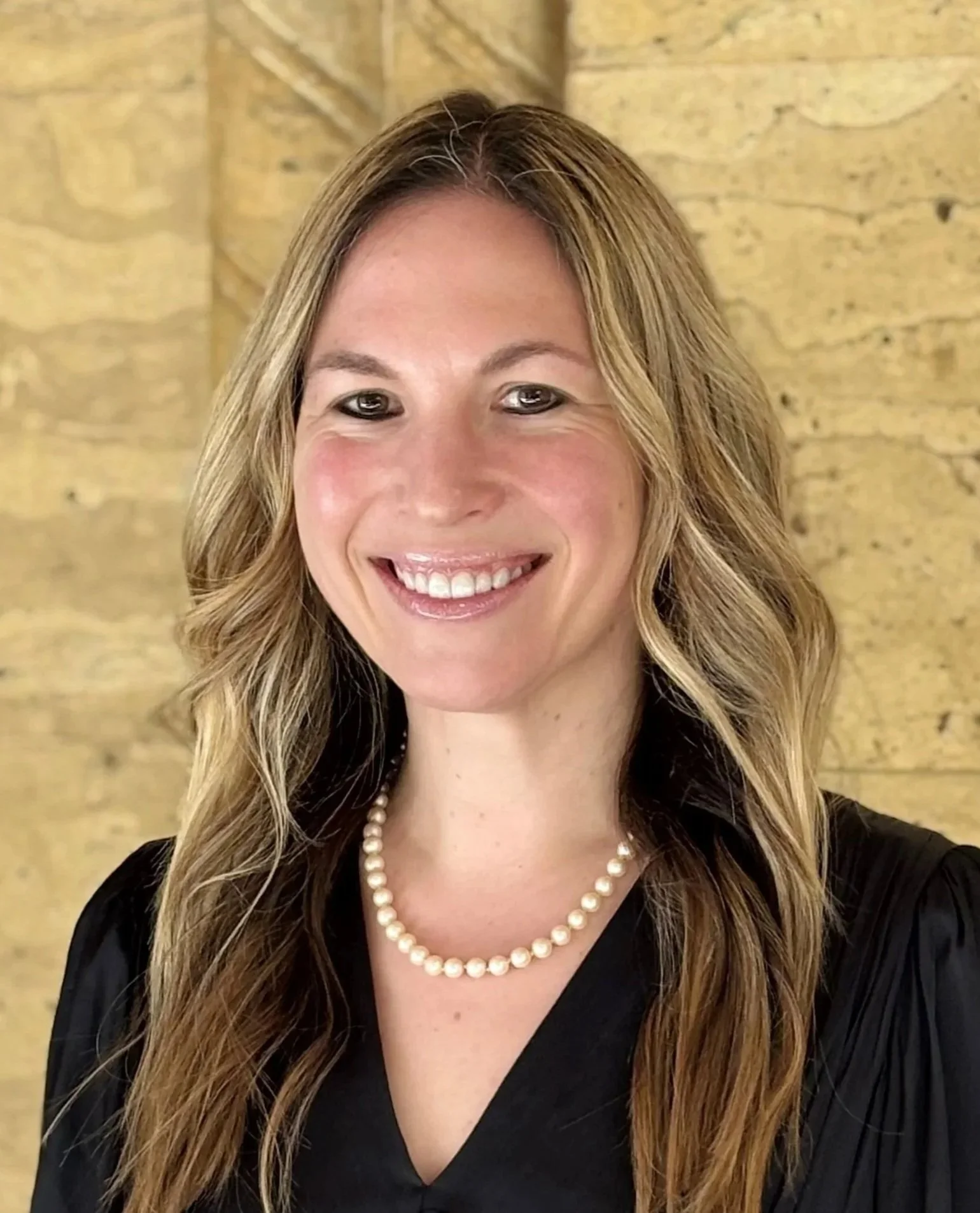 A smiling woman with long wavy blonde hair, wearing a pearl necklace and a black top, standing in front of a beige stone wall.