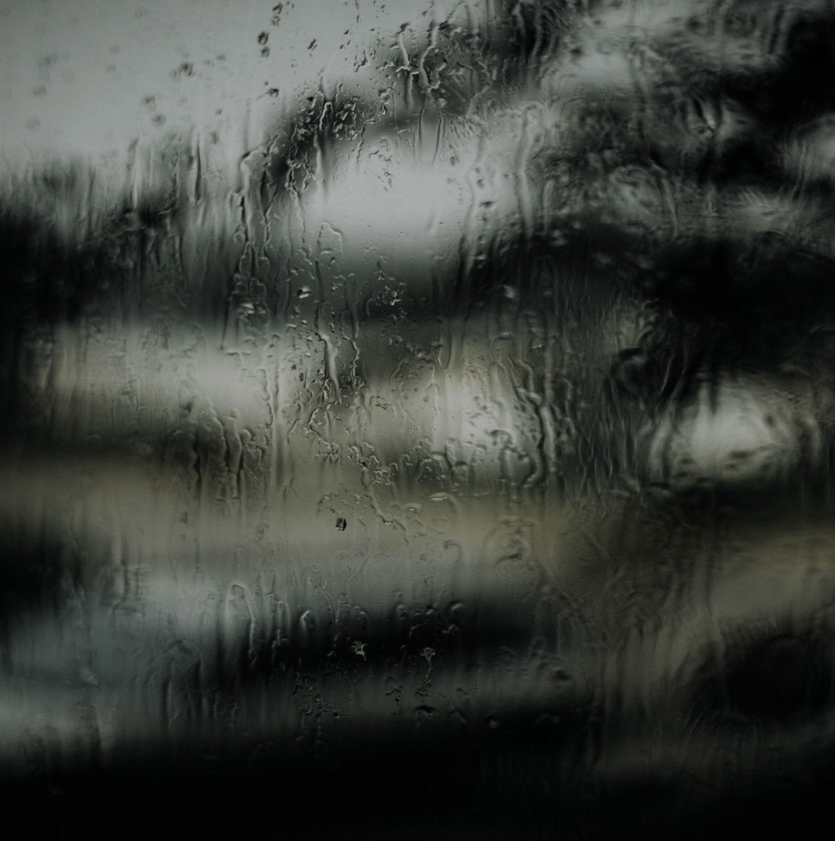 A view through a rain-covered window showing a blurry, gray, and overcast outdoor scene with trees and buildings, streaked and blurred by the rain on the glass.