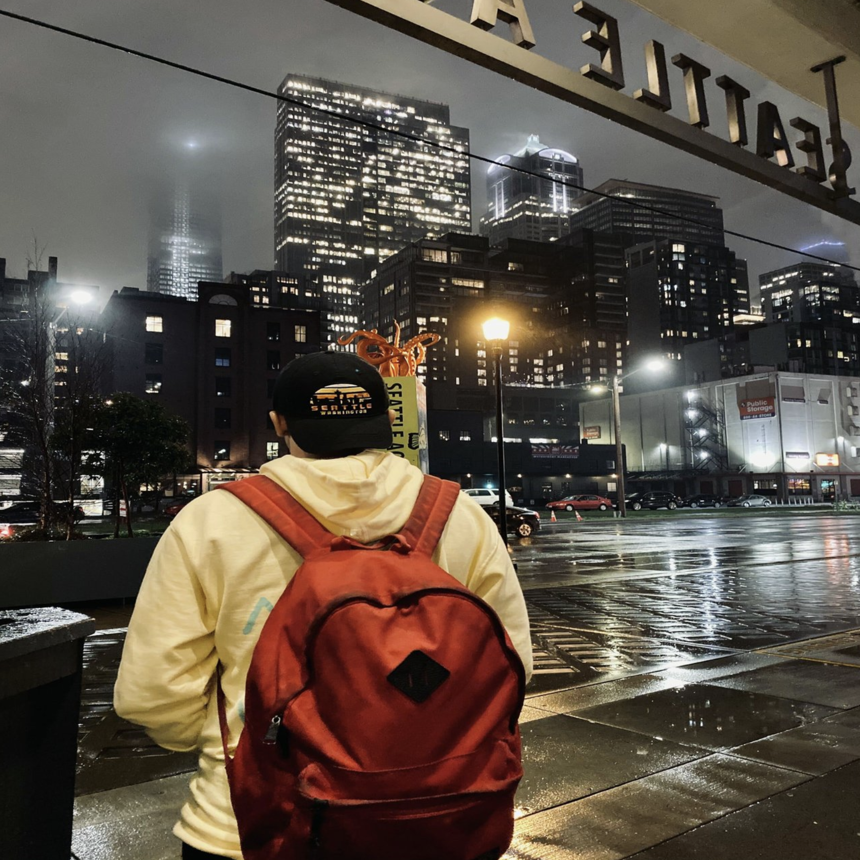 A person with a red backpack and a black cap stands on a rainy city street at night, overlooking illuminated skyscrapers in a foggy urban scene.