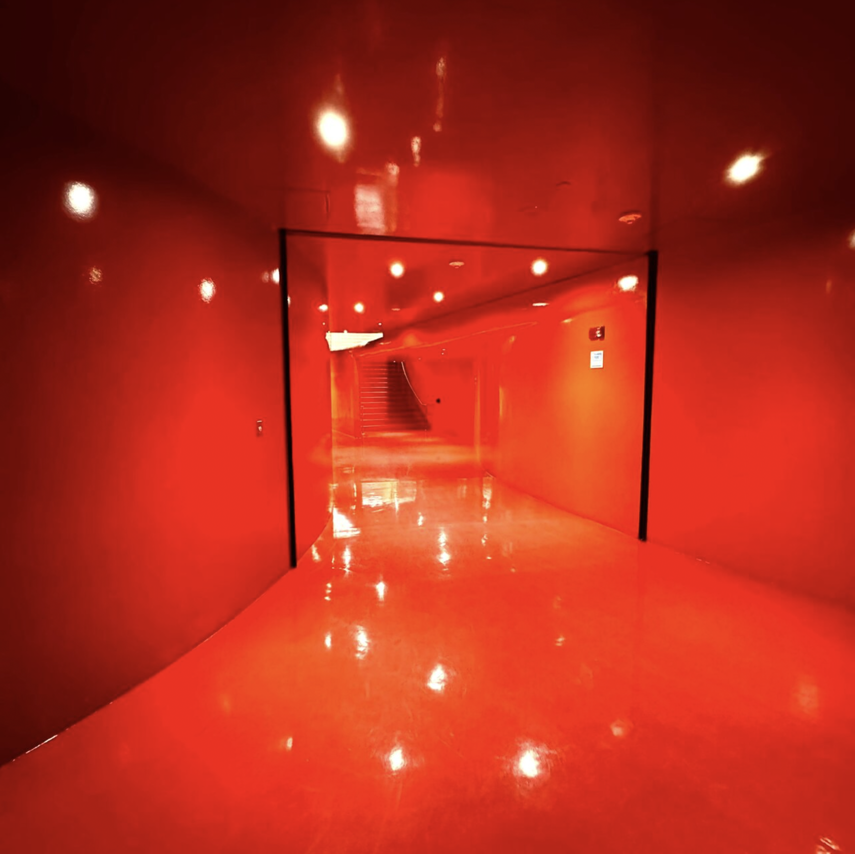 A vivid, red atmospheric hallway with reflective polished flooring, bright ceiling lights, and a staircase at the end.