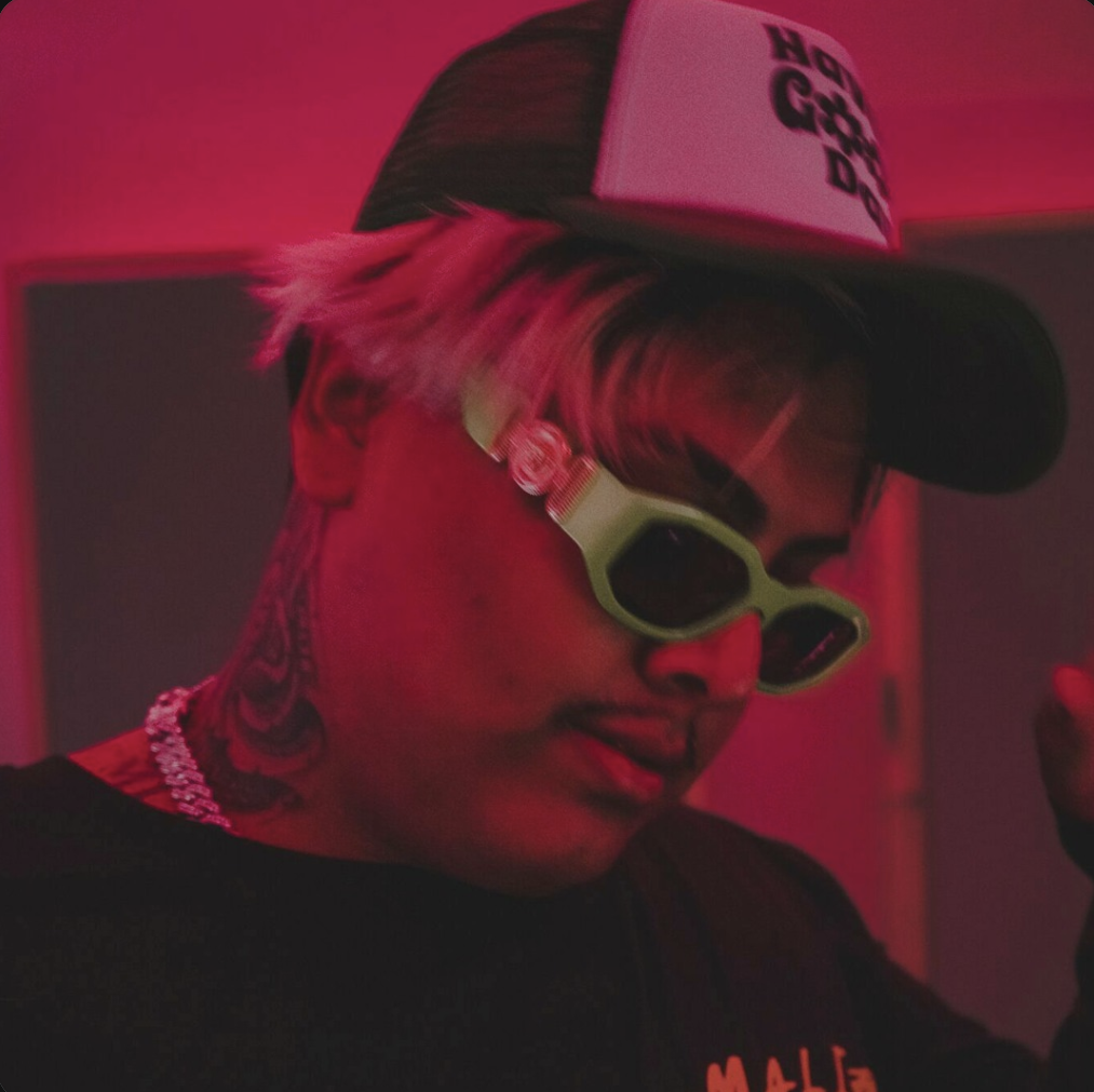 Person with tattoos on neck, wearing green sunglasses, a black cap, and a white cap with black text, gesturing with hand in a dimly lit room with pink/red lighting.
