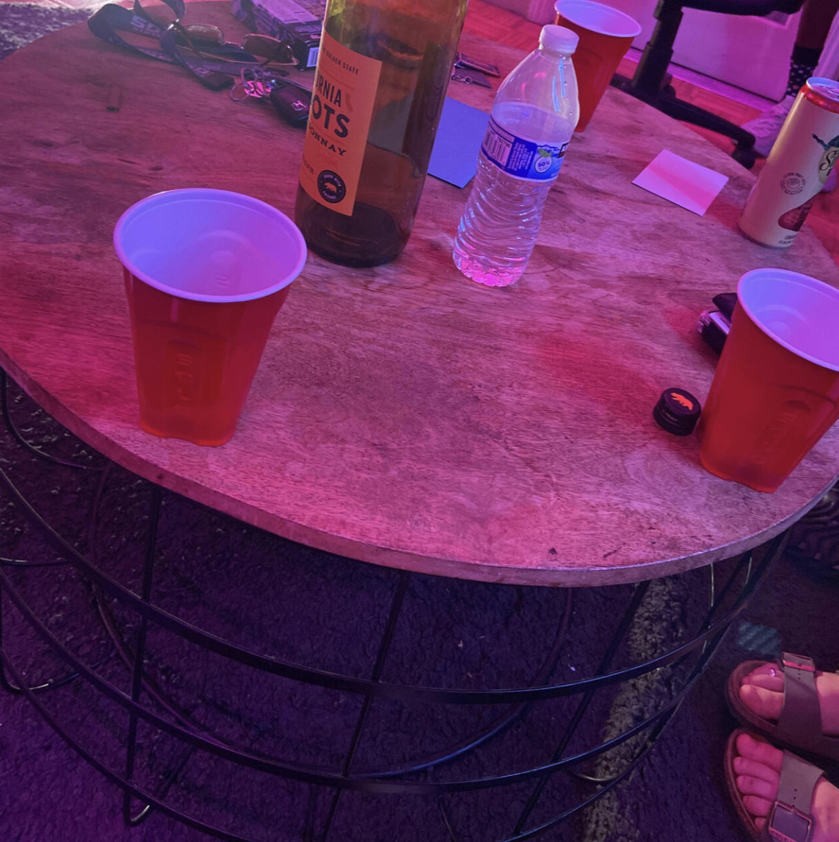 A round wooden table with two red plastic cups, an empty water bottle, a beer bottle, a red cup, and various personal items in a dimly lit setting with purple lighting.