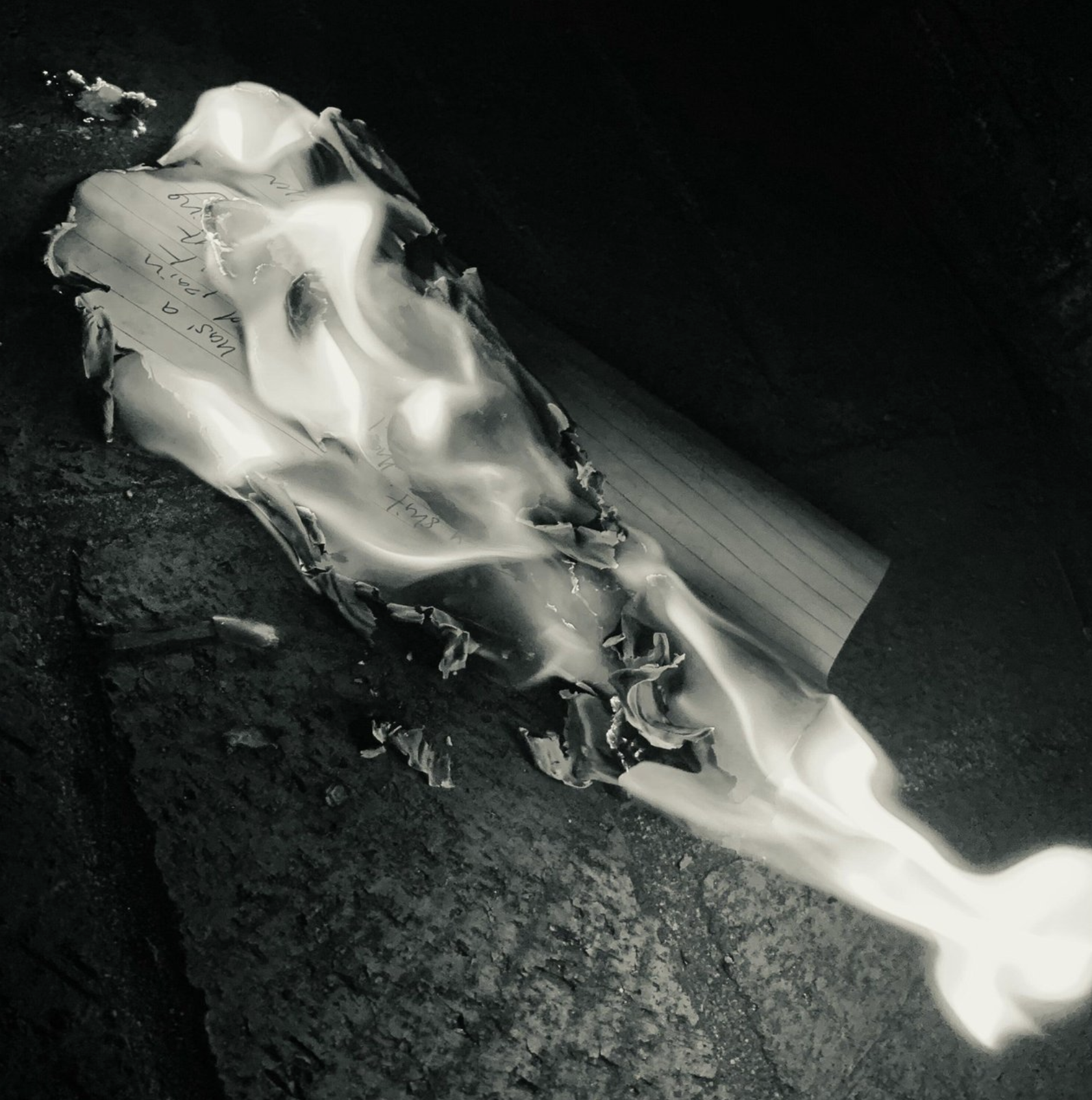 Burned piece of crumpled paper with handwritten notes and a lined index card attached, lying on a rough black surface.