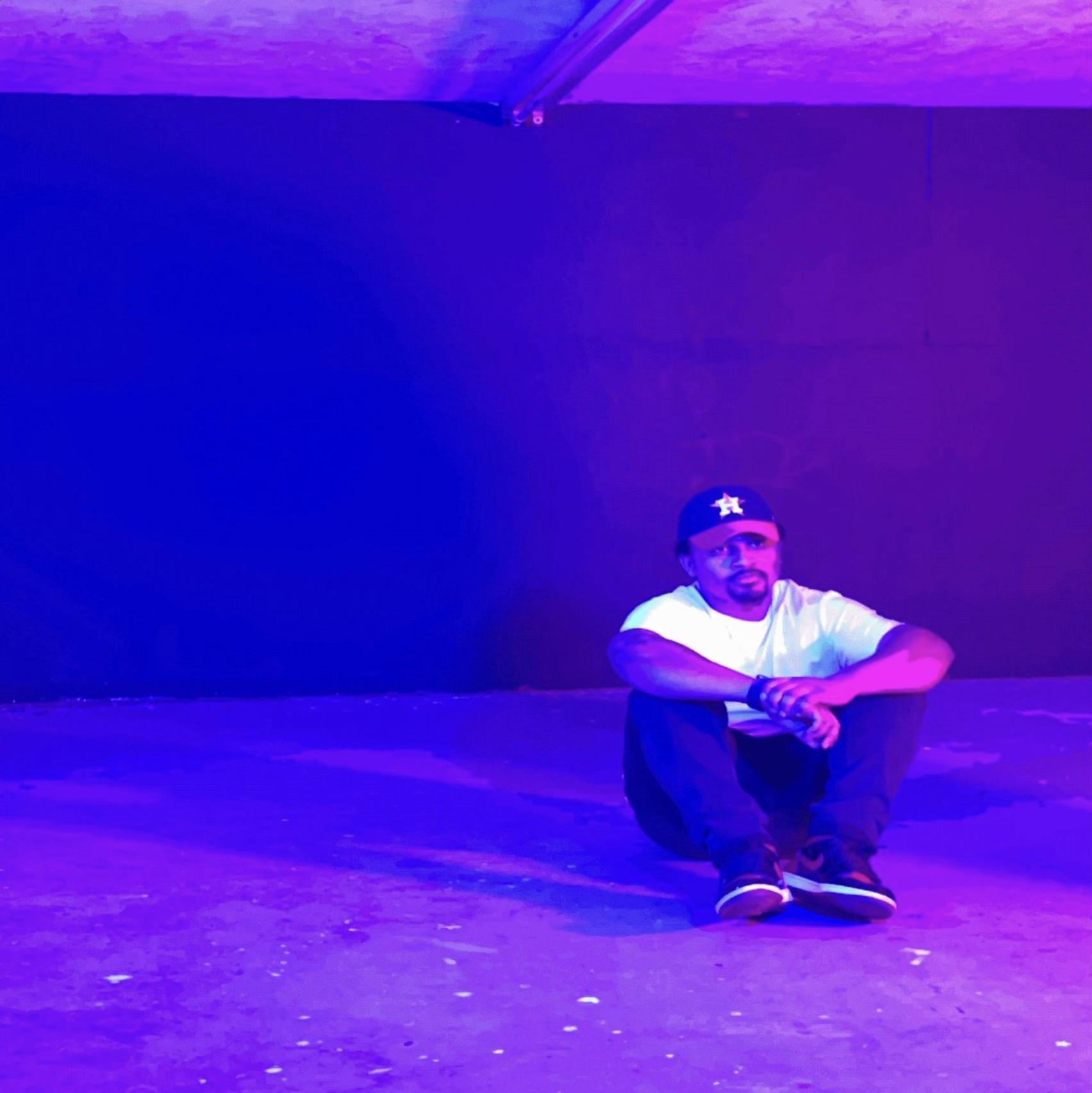 A man sitting on the floor with purple and blue lighting, wearing a white t-shirt, black pants, and a black cap with a star symbol, against a dark background.