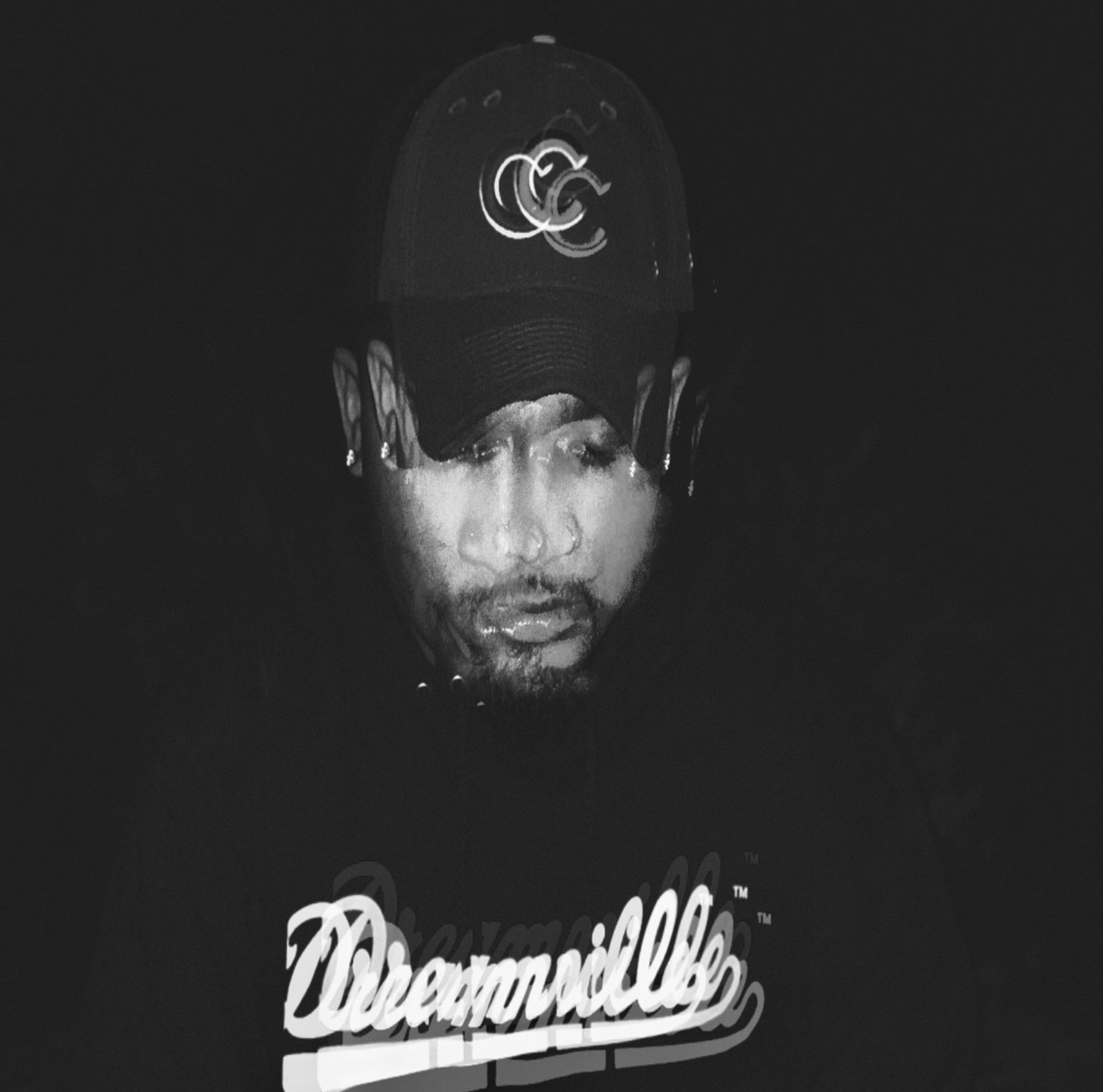 A person wearing a baseball cap with a logo, partially submerged in darkness, with their face and upper torso visible, wearing a sweatshirt with the word 'Decamillle' on it.