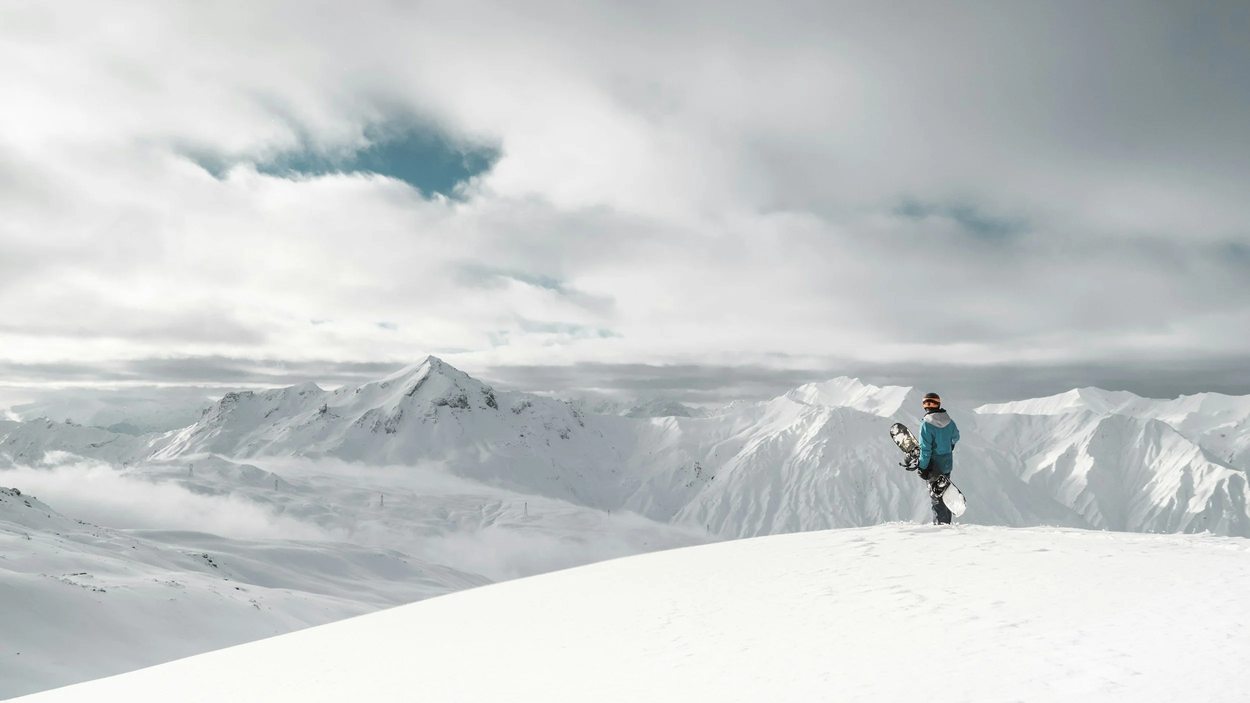 A person in a blue jacket and snowboard boots holding a snowboard stands on snow-covered terrain in a mountainous landscape with snow-capped peaks and cloudy sky.