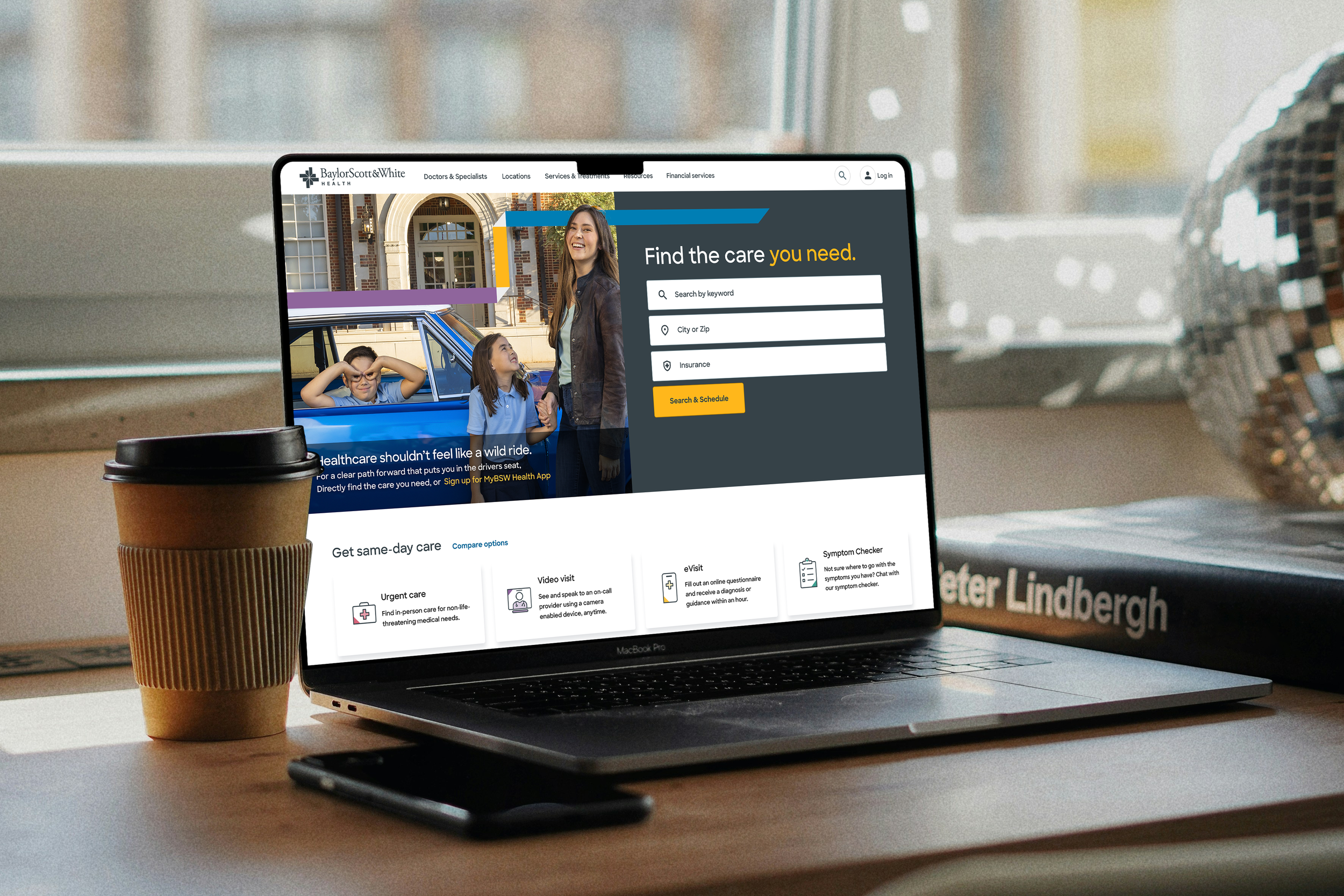 Open laptop on a desk displaying a healthcare website with a woman and children, a coffee cup, a smartphone.