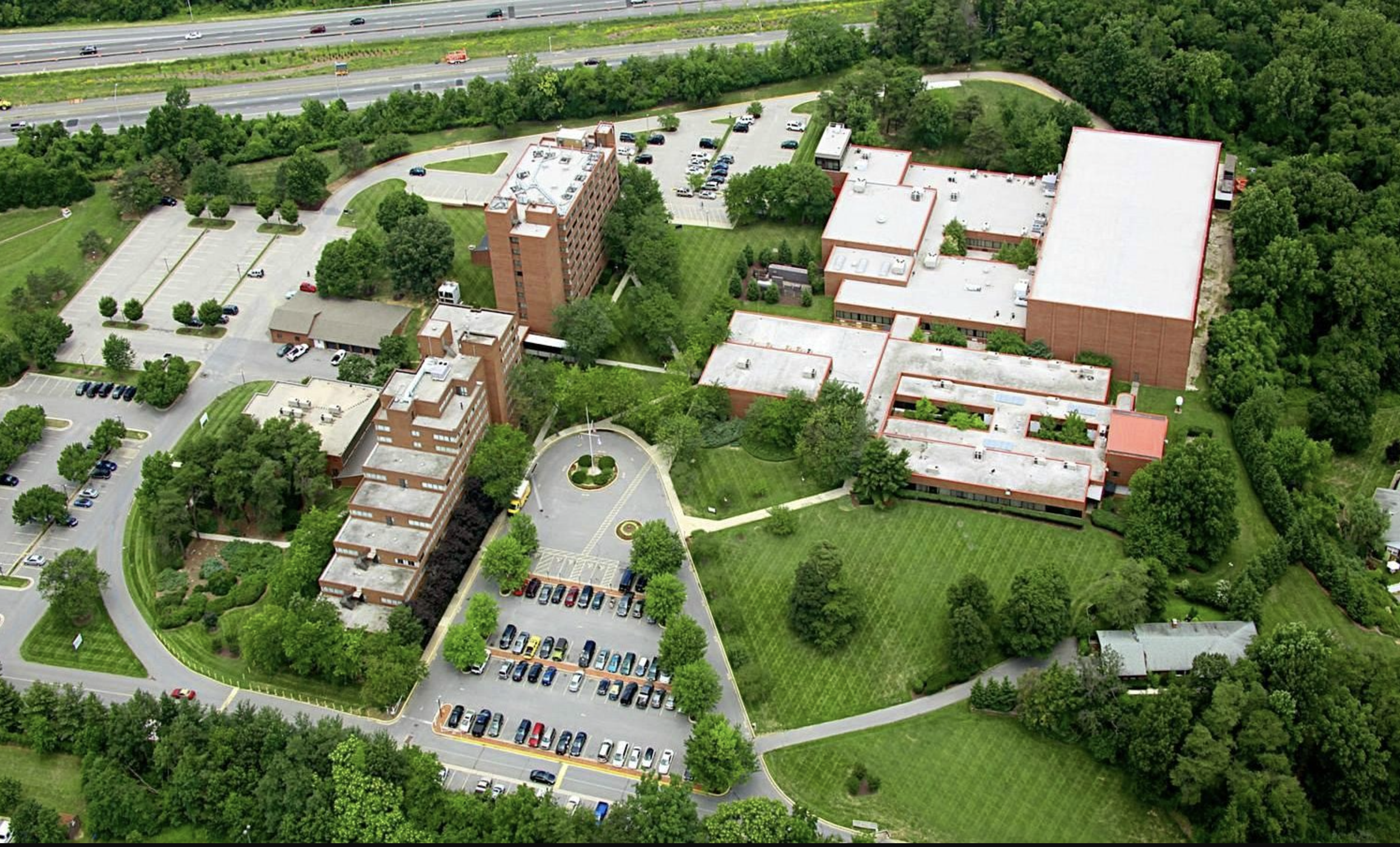 Aerial view of a multi-building campus surrounded by green trees, parking lots, and a nearby highway with traffic.