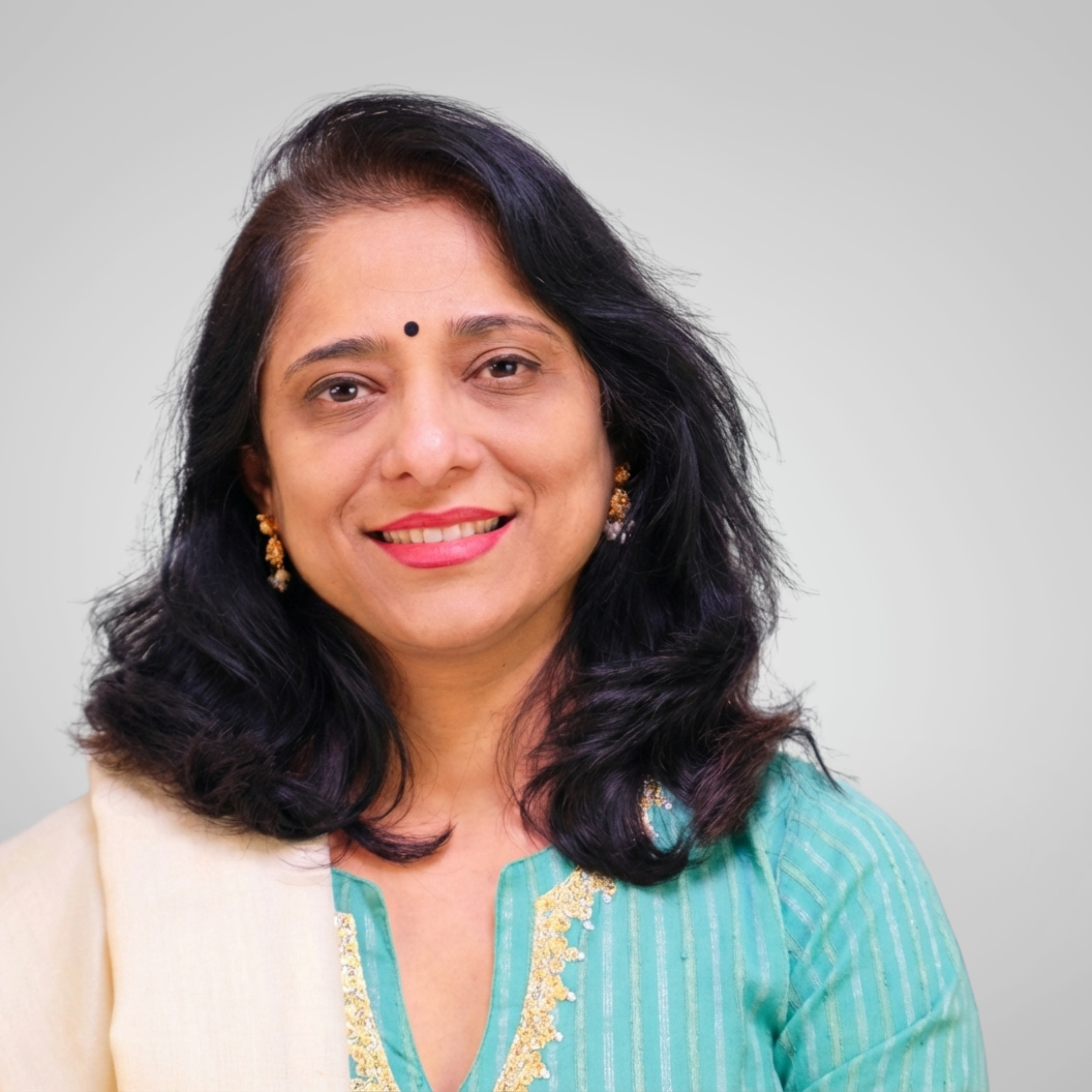A professional headshot of a smiling woman with black hair, wearing traditional Indian attire with gold earrings.