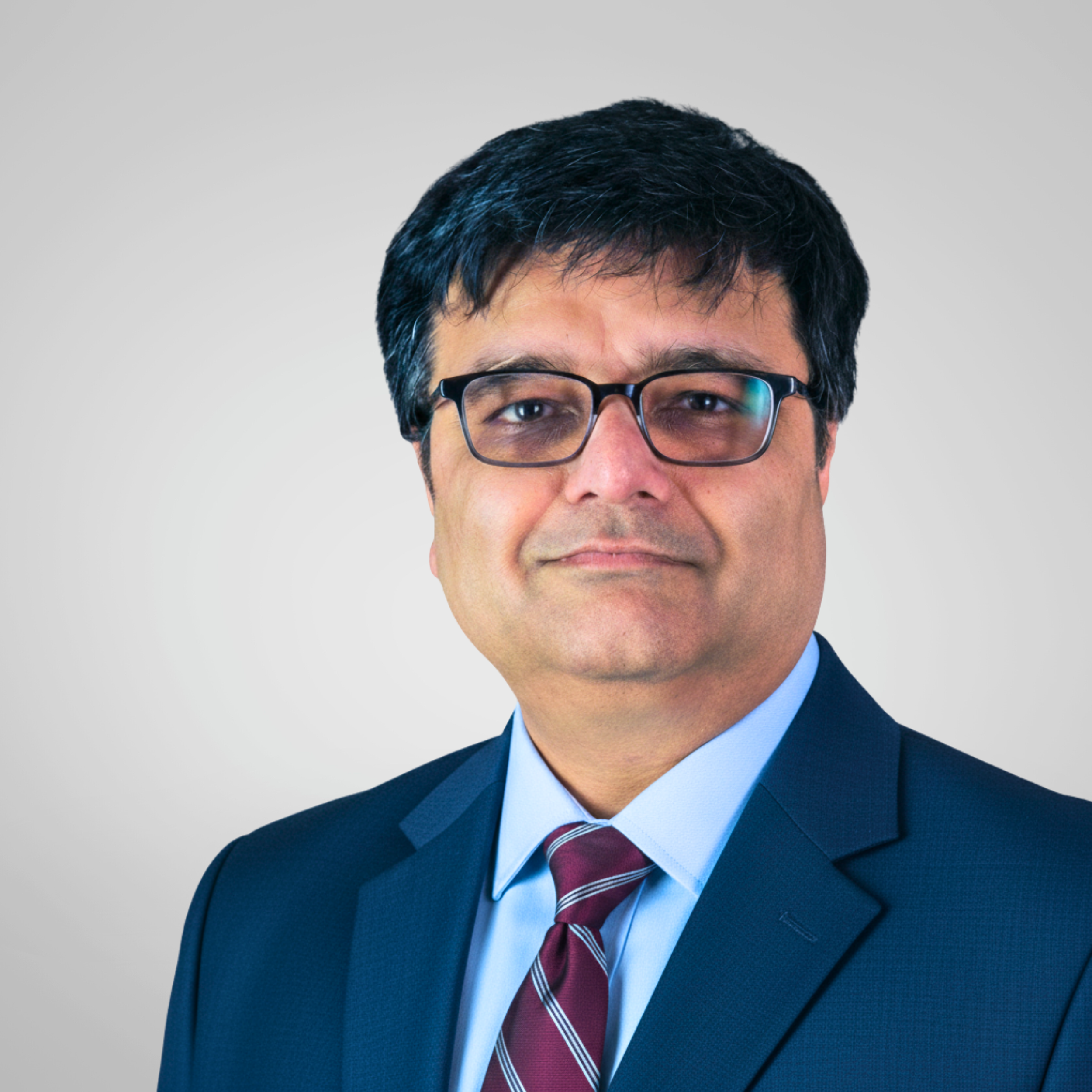 Portrait of a man with glasses wearing a dark pinstripe suit, yellow shirt, and red patterned tie, standing in front of a textured beige and orange background.