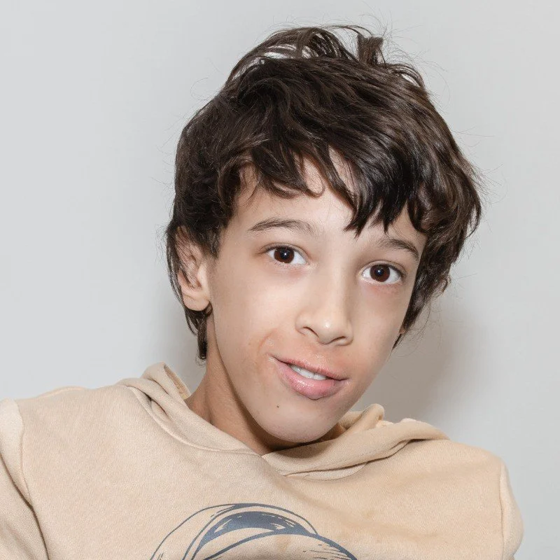 A young person with short, dark brown, curly hair and light skin, wearing a beige hoodie, standing against a plain white background.