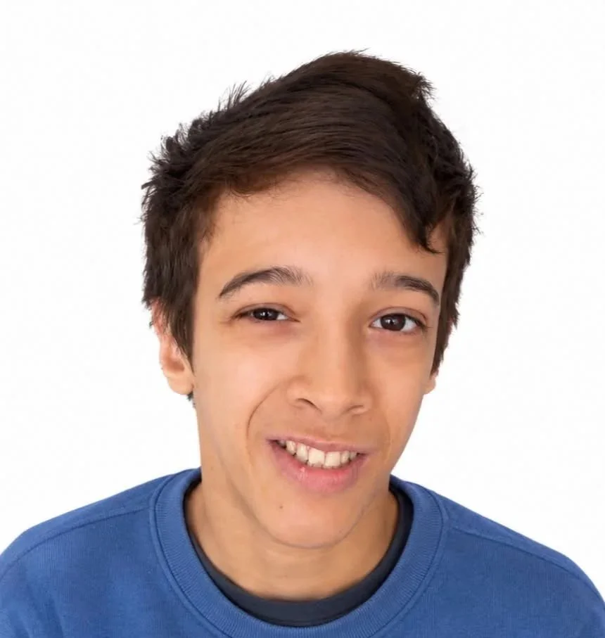 A young man with brown hair wearing a blue shirt, smiling with one eye slightly squinted against a plain white background.