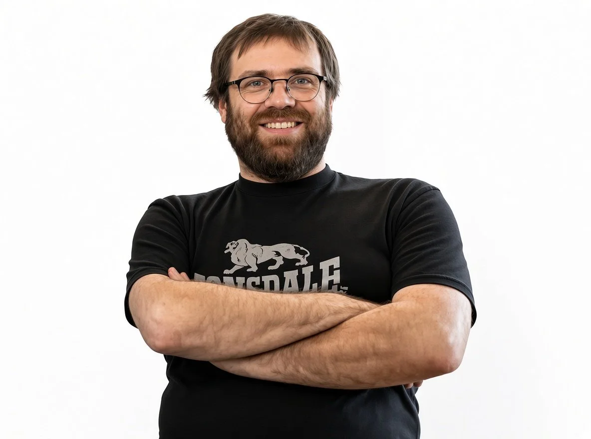 A man with glasses and a beard, wearing a black T-shirt with a lion graphic and crossed arms, smiling at the camera against a white background.