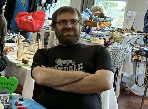 A man with glasses and a beard, wearing a black T-shirt with a graphic and crossed arms, sitting at a table in an indoor setting with tables, items, and people in the background.