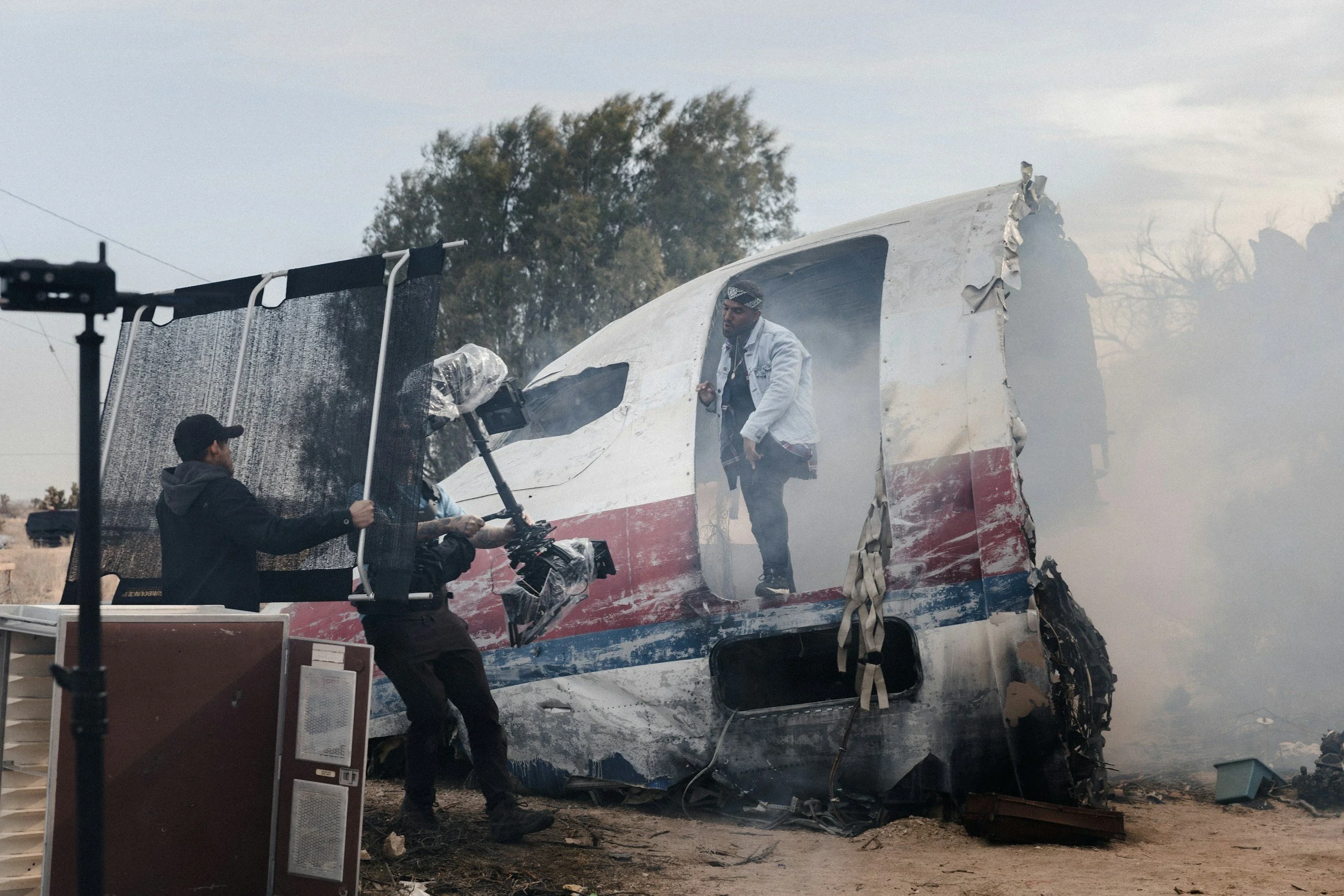 Film crew on set with damaged airplane propeller and body, preparing for a scene, with a man walking out of the wreckage, surrounded by equipment and smoke.