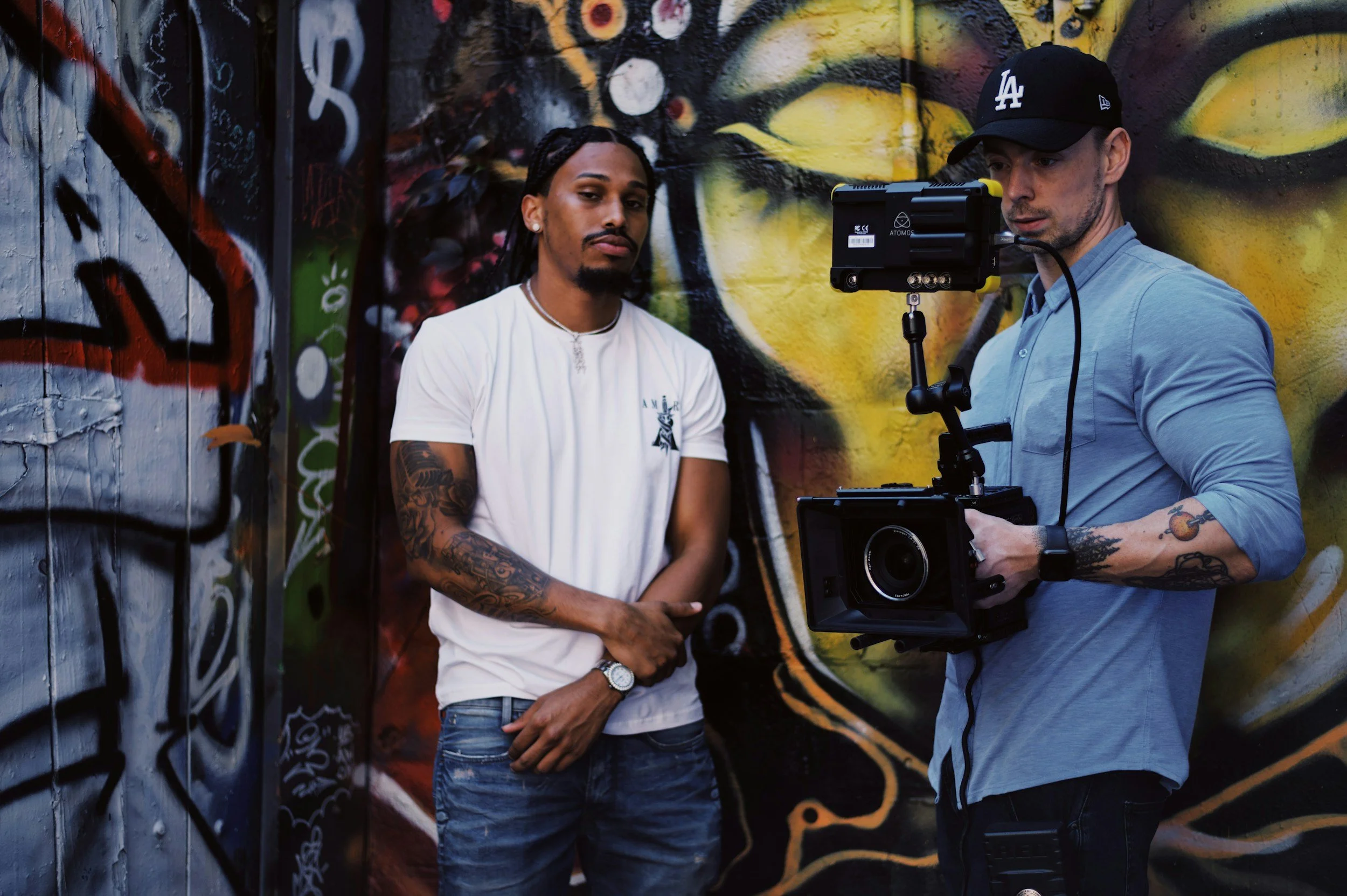 Two men standing in front of a colorful graffiti wall. One man is wearing a white t-shirt, has tattoos on his arms, and looks at the camera. The other man is wearing a light blue long sleeve shirt and a black LA baseball cap, and is handling filming equipment.