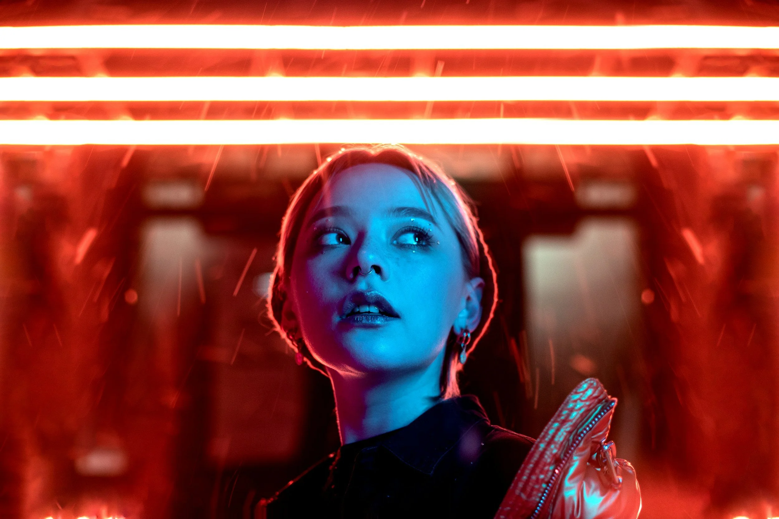 A young woman with short hair and earrings looking upward in front of red neon lights.