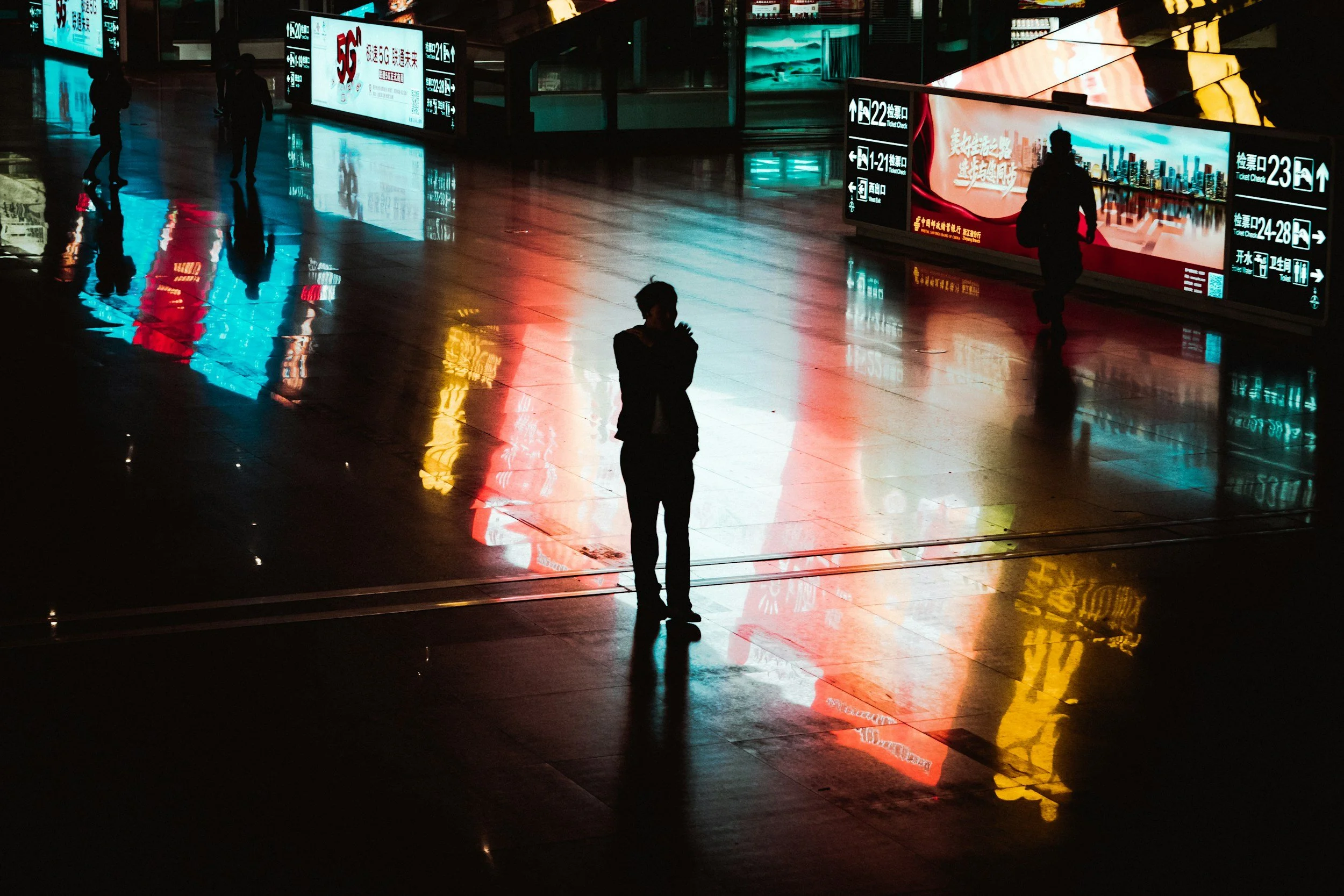 Silhouettes of four people walking inside a shopping mall at night, with vibrant reflections on the polished floor and illuminated advertisements in the background.