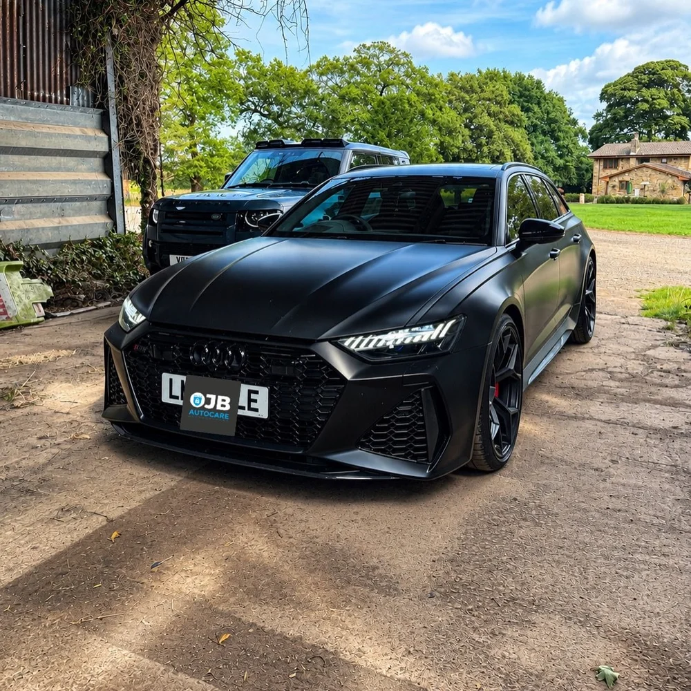 Satin black Audi RS6 parked outside with an OJB badge on the number plate and a Land Rover behind it
