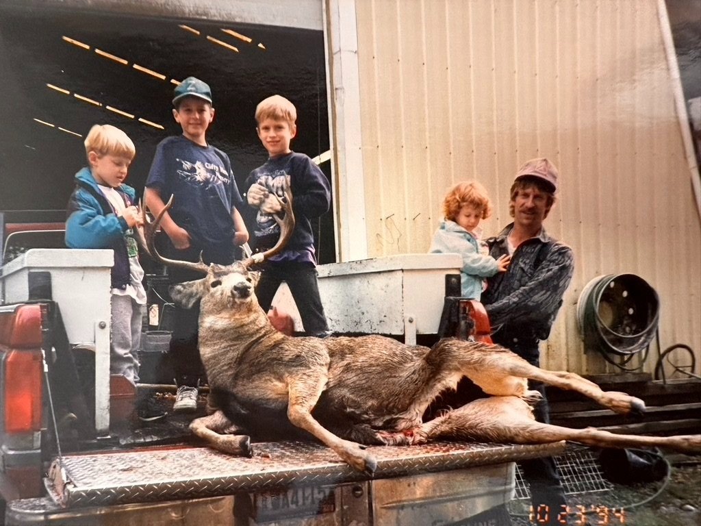 A man and four children standing in the back of a pickup truck with a large deer they have hunted, with one of the children holding the deer's antlers.