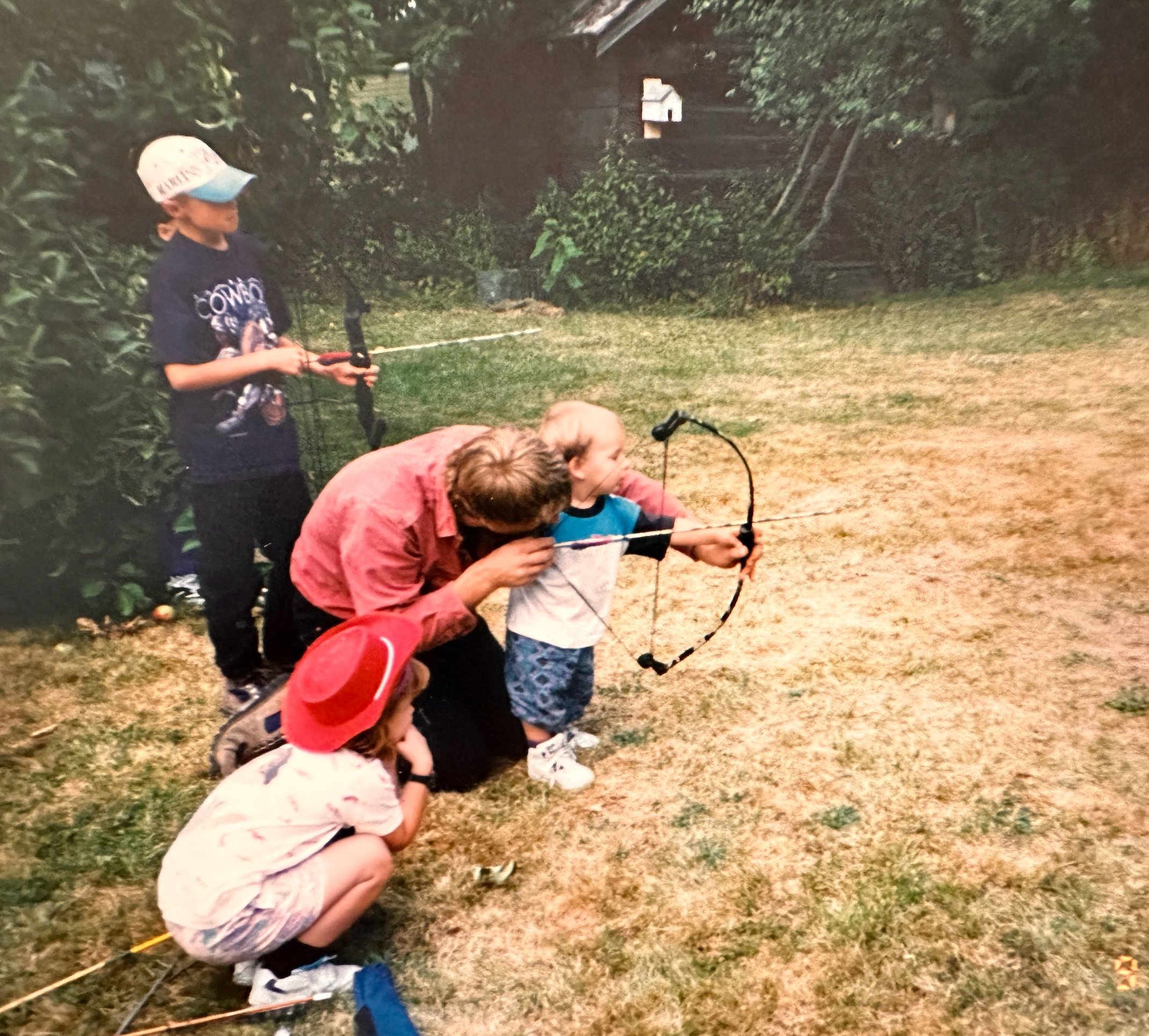 Two children and an adult outdoors, practicing archery, with one child aiming a bow and the adult assisting. A girl kneels in the foreground wearing a red hat, a boy in the background is holding a bow with an adult helping him, and another boy stands