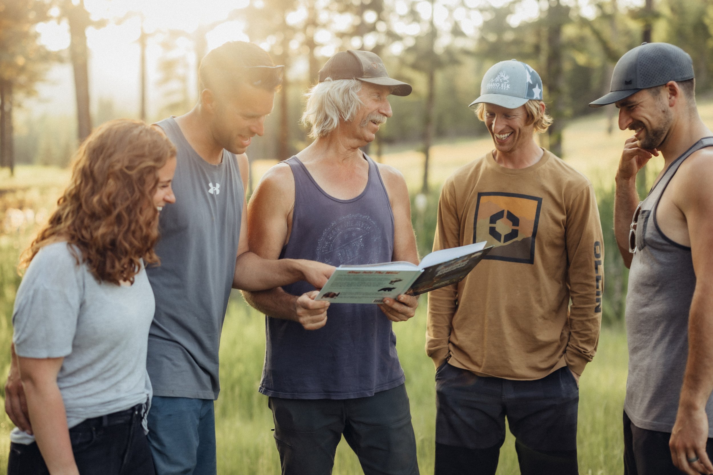 Group of six smiling people standing outdoors in a forest, looking at a brochure.